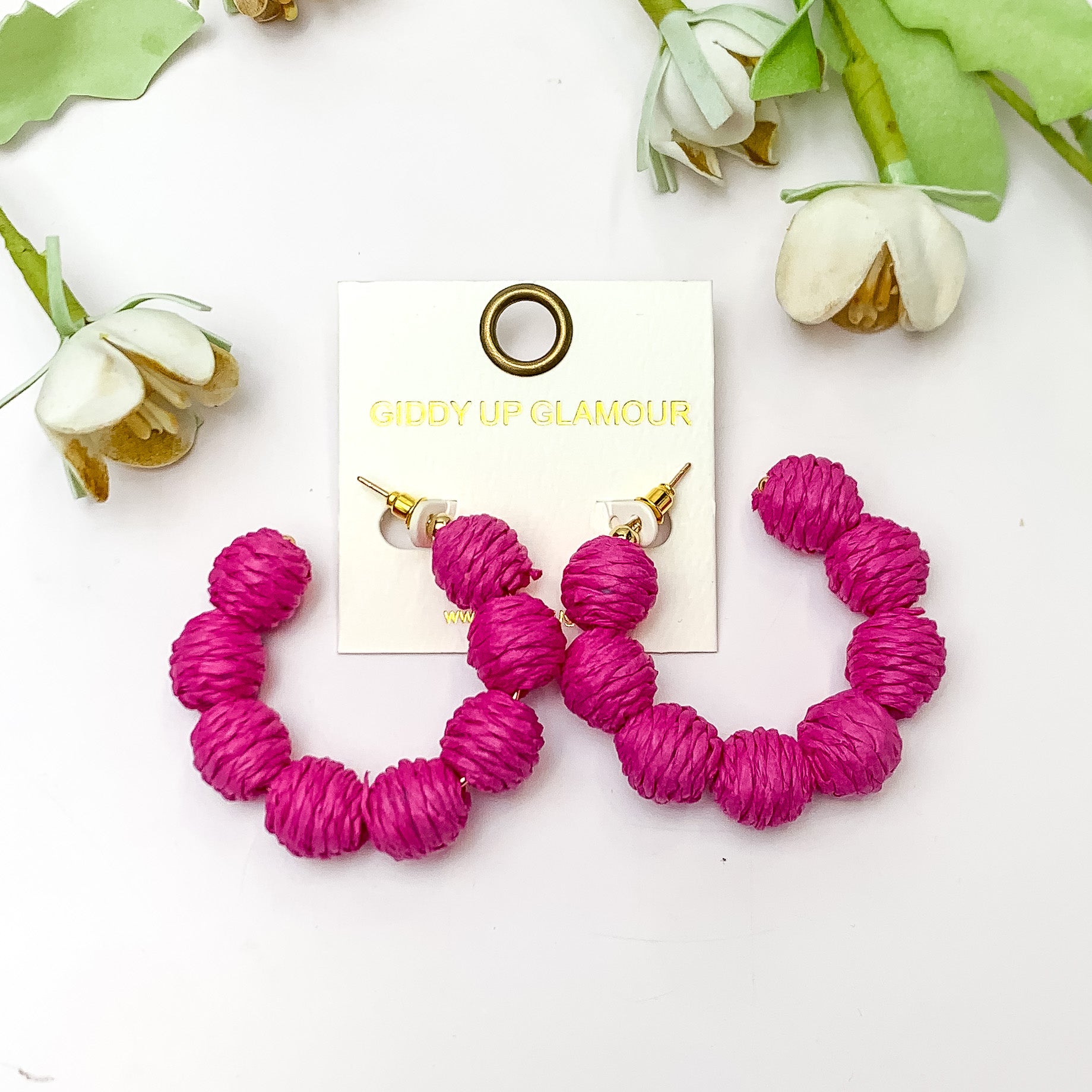 Sorbet Summer Raffia Ball Hoop Earrings in Hot Pink. Pictured on a white background with flowers above the earrings.