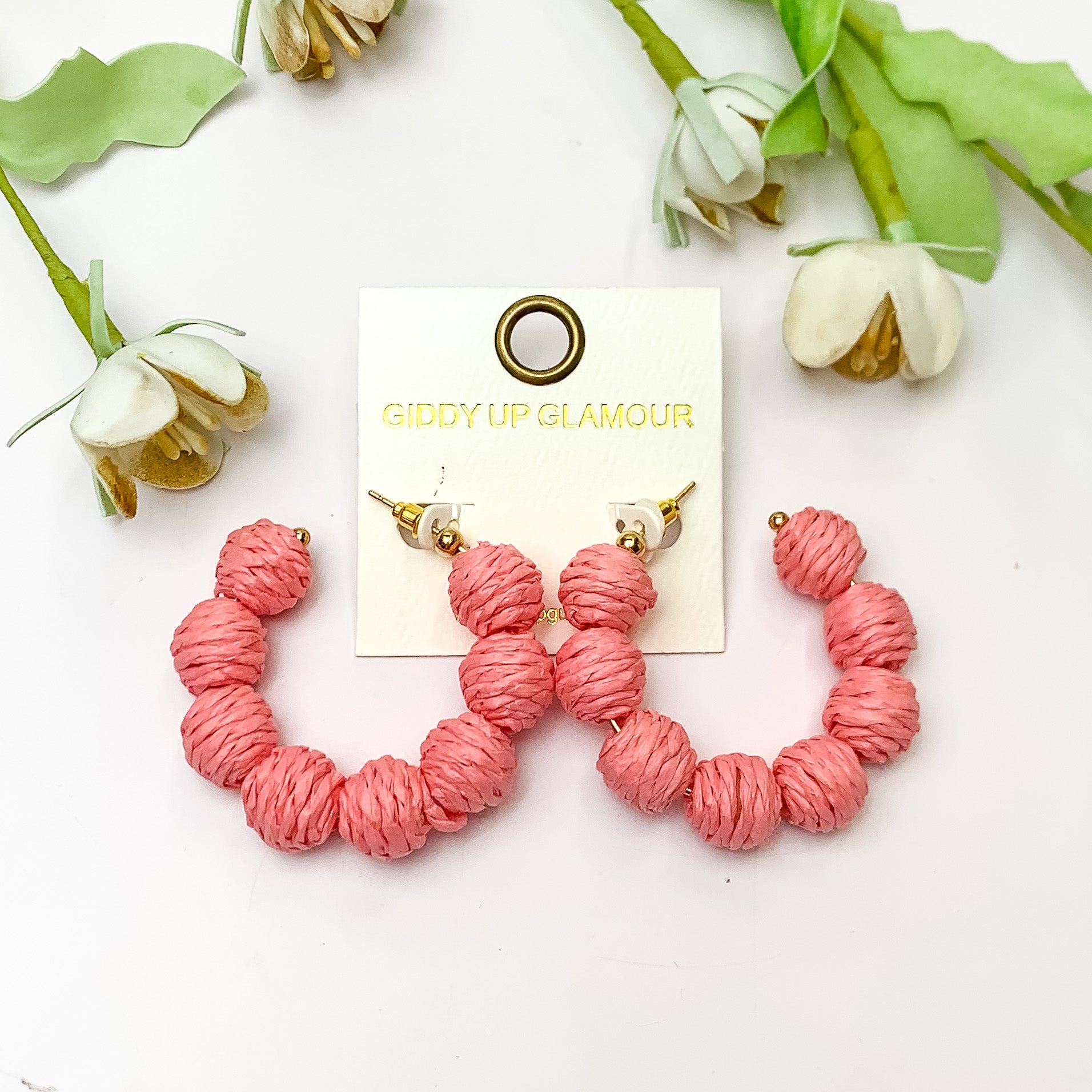 Sorbet Summer Raffia Ball Hoop Earrings in Pink. Pictured on a white background with flowers above the earrings.