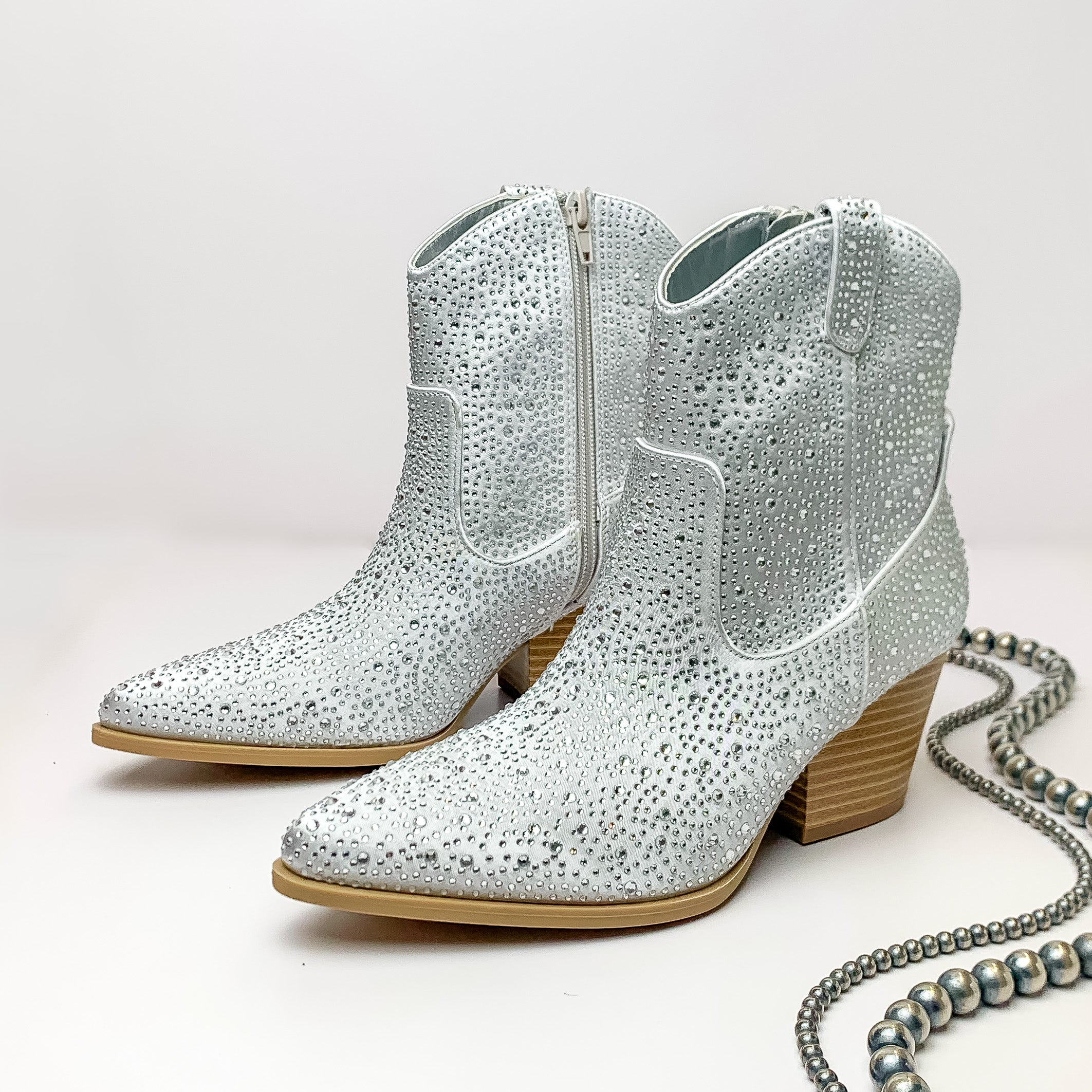 Nashville Sparkle Heeled Ankle Booties with Clear Crystals in Silver. Pictured on a white background with jewelry laying on the platform. 