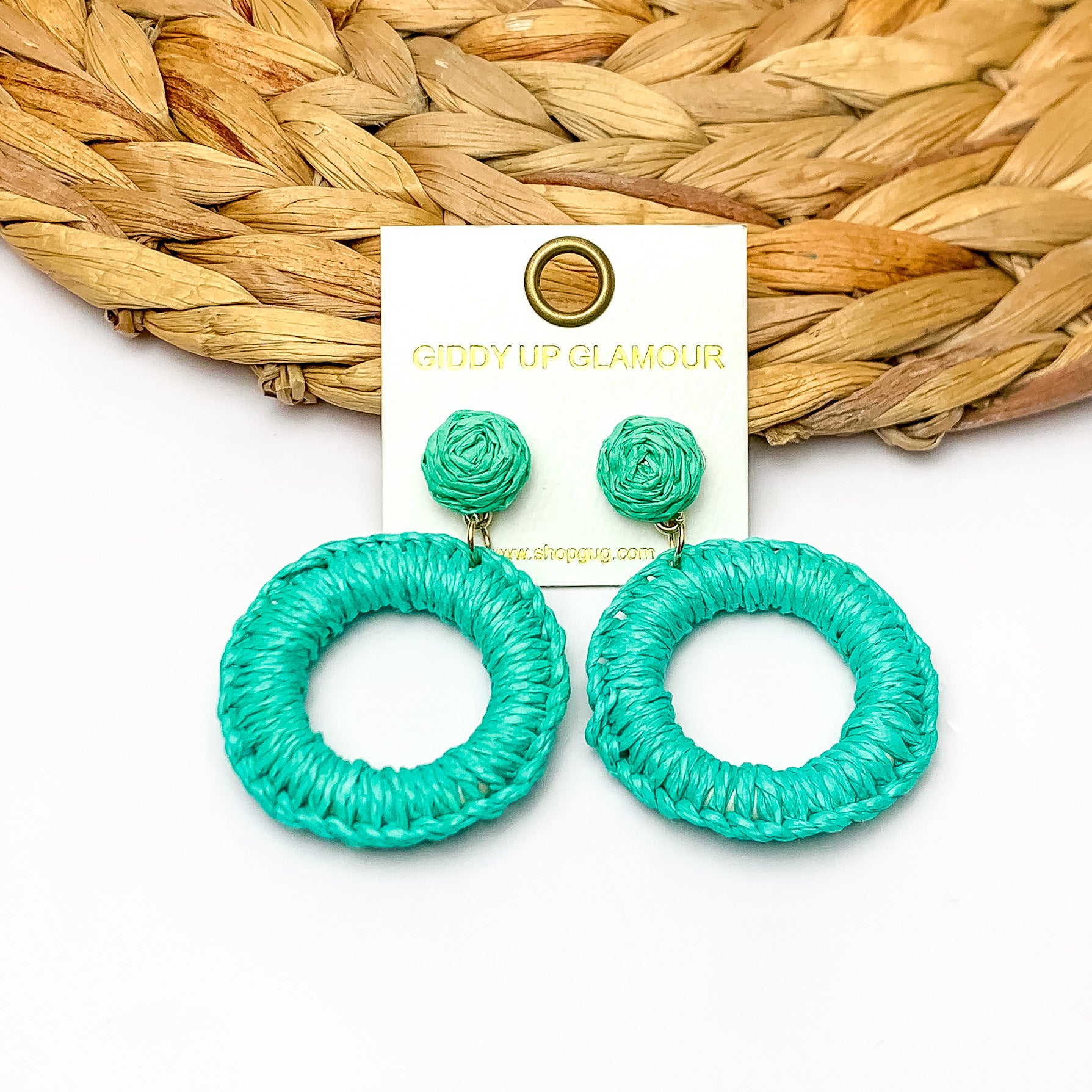 Beachside Café Raffia Wrapped Circle Earrings in Turquoise Green. Pictured on a white background with the top of the earrings laying on a brown circle decorative piece.