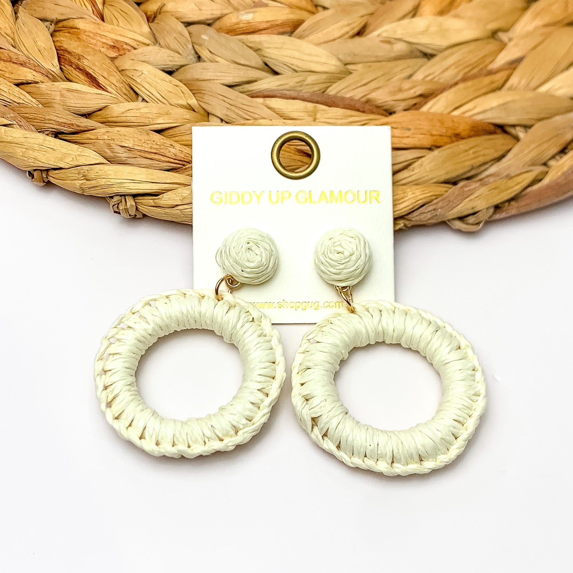 Beachside Café Raffia Wrapped Circle Earrings in Ivory White. Pictured on a white background with the top of the earrings laying on a brown circle decorative piece.