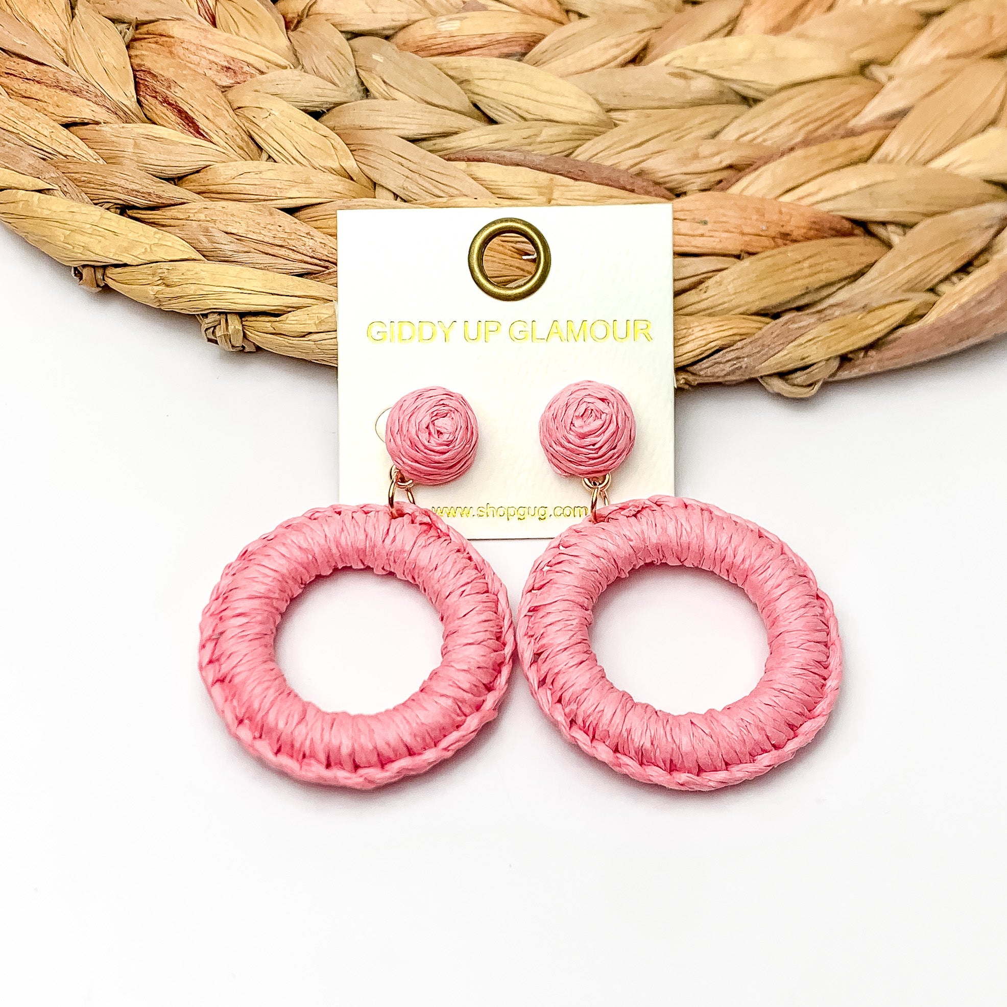 Beachside Café Raffia Wrapped Circle Earrings in Light pink. Pictured on a white background with the top of the earrings laying on a brown circle decorative piece.