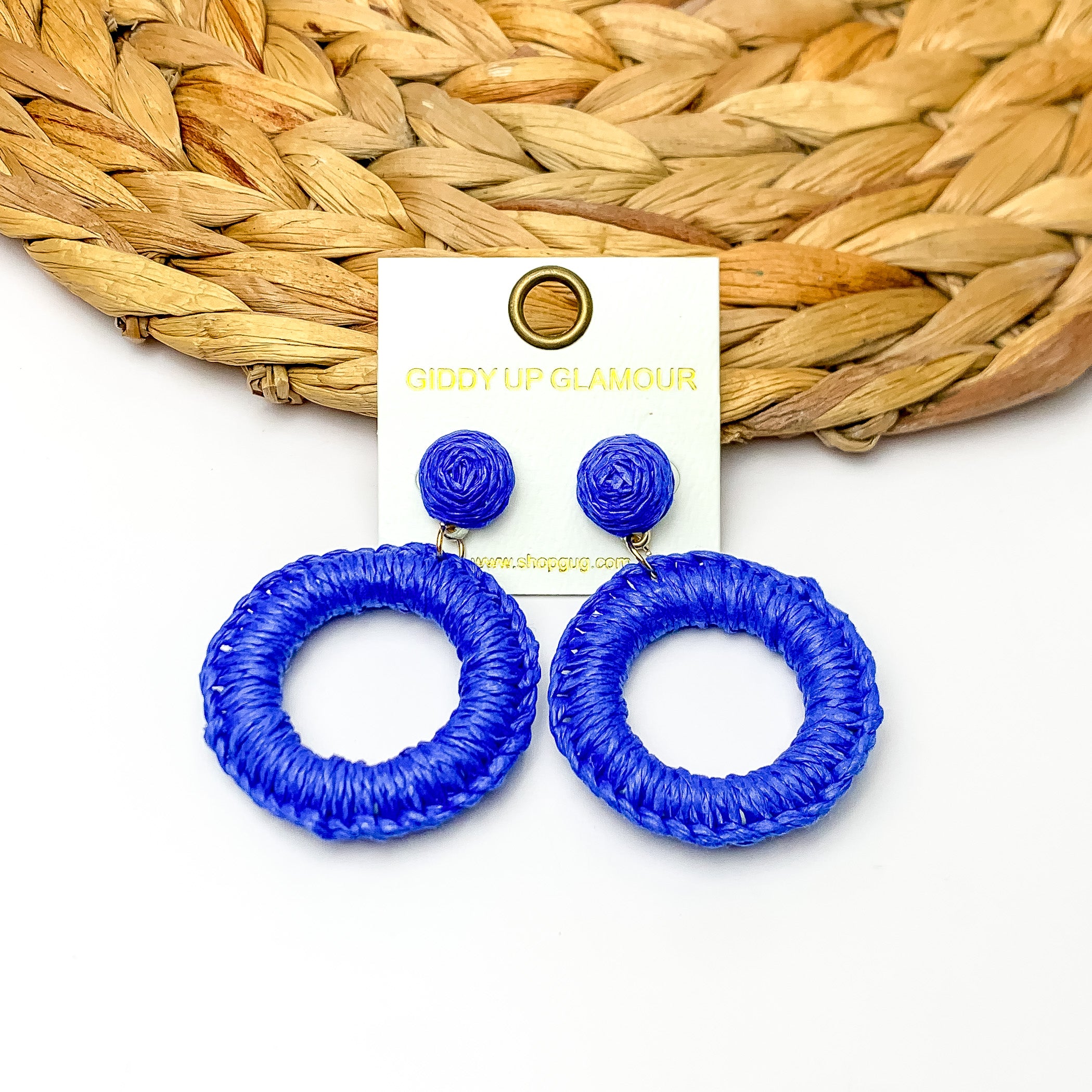 Beachside Café Raffia Wrapped Circle Earrings in Royal Blue. Pictured on a white background with the top of the earrings laying on a brown circle decorative piece. 