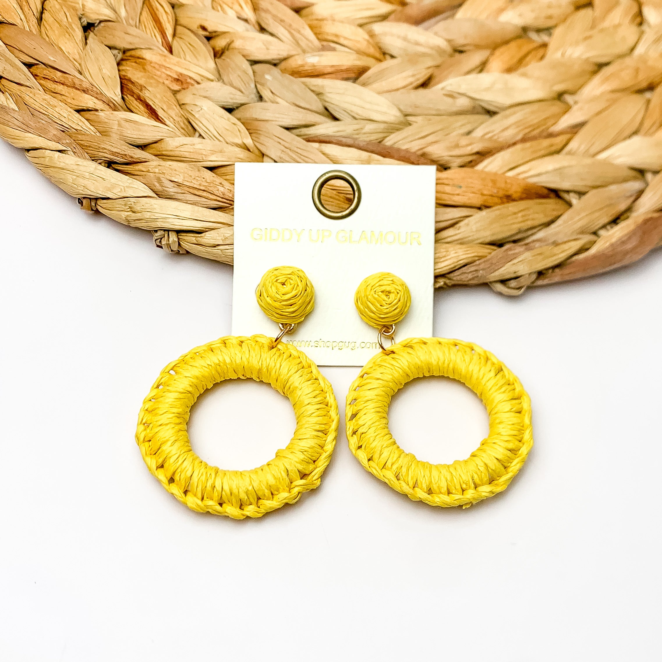 Beachside Café Raffia Wrapped Circle Earrings in Yellow. Pictured on a white background with the top of the earrings laying on a brown circle decorative piece.