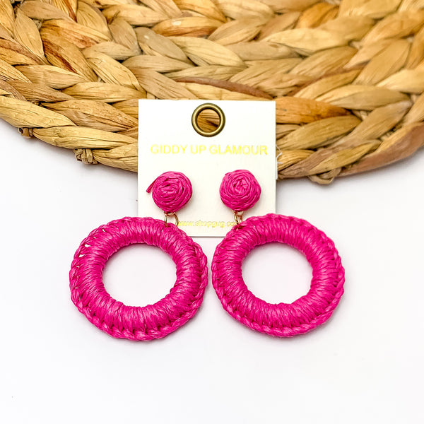 Beachside Café Raffia Wrapped Circle Earrings in Fuchsia Pink. Pictured on a white background with the top of the earrings laying on a brown circle decorative piece.