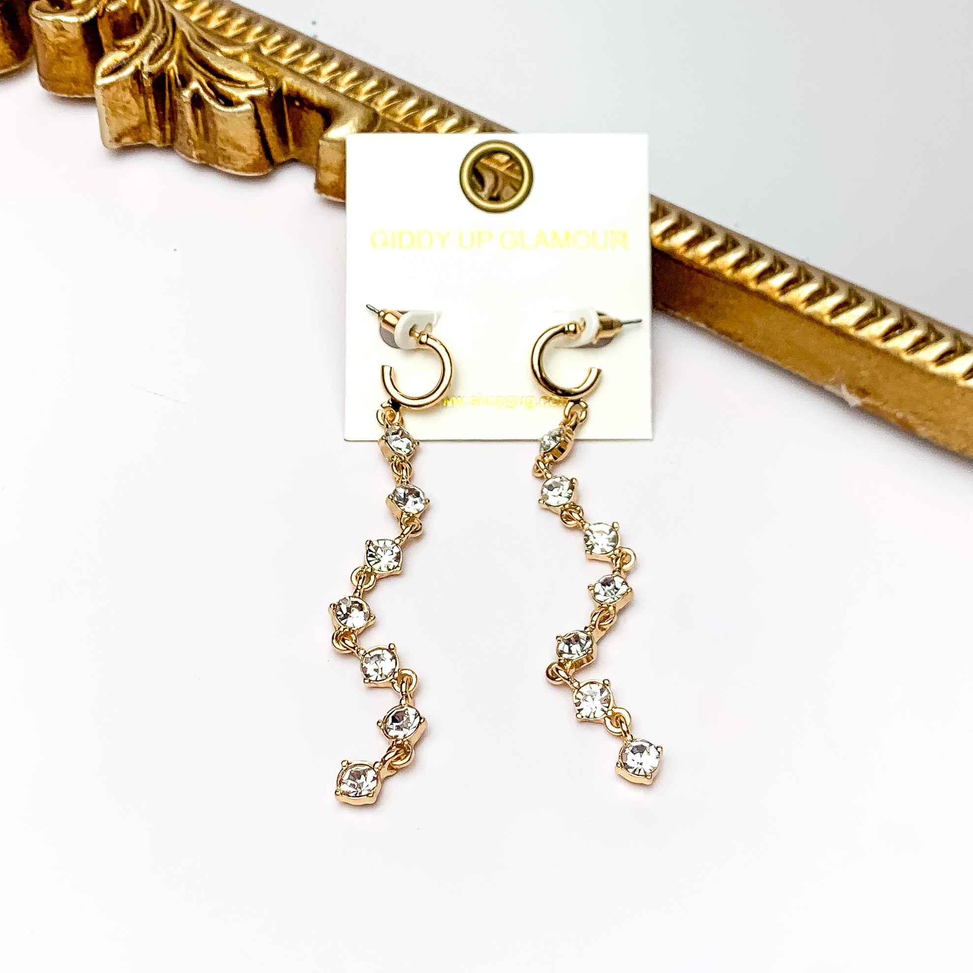 Red Carpet Moment Long Gold Tone Earrings With Clear Crystals. Pictured on a white background with a gold frame behind the earrings.