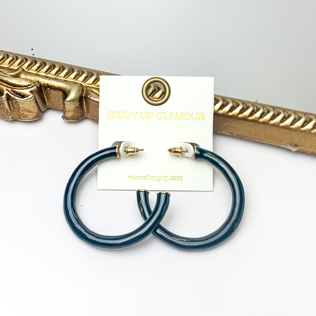 Plan For Cabo Large Hoop Earrings in Black. Pictured on a white background with a gold frame behind the earrings.