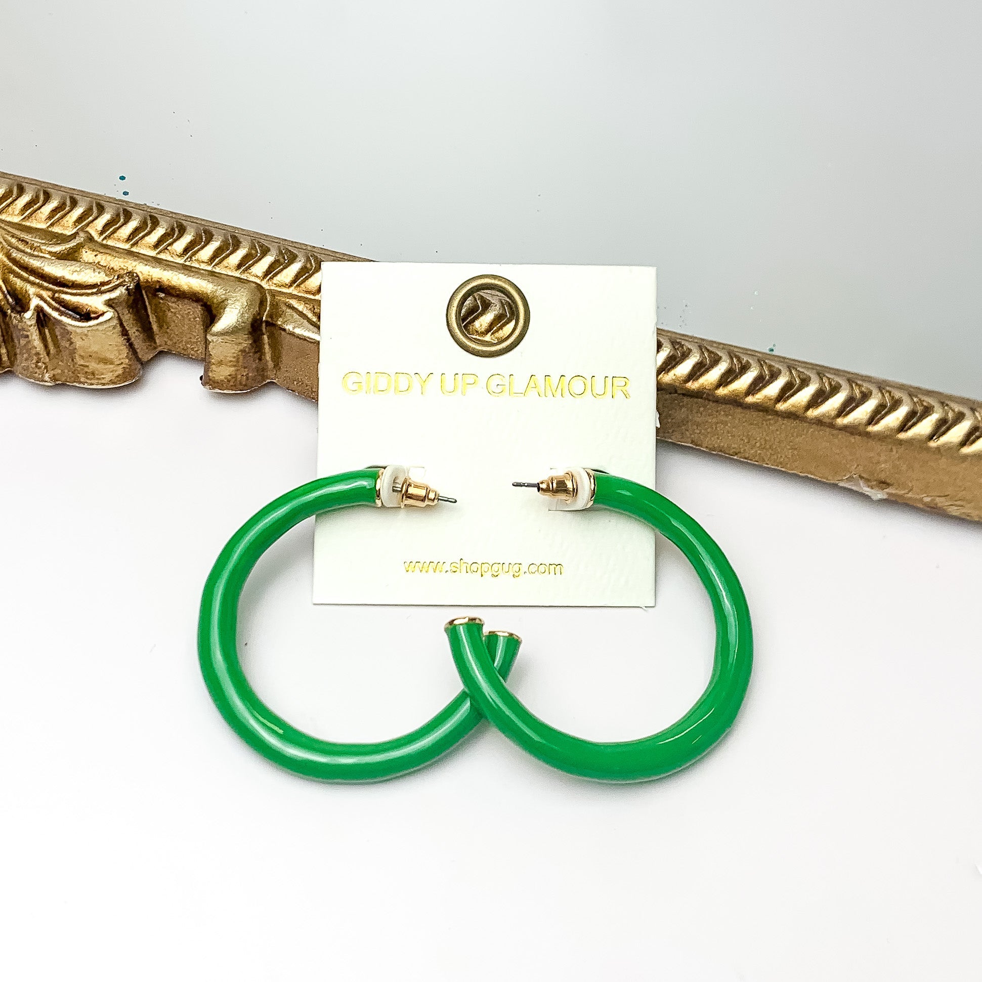 Plan For Cabo Large Hoop Earrings in Green. Pictured on a white background with a gold frame behind the earrings.