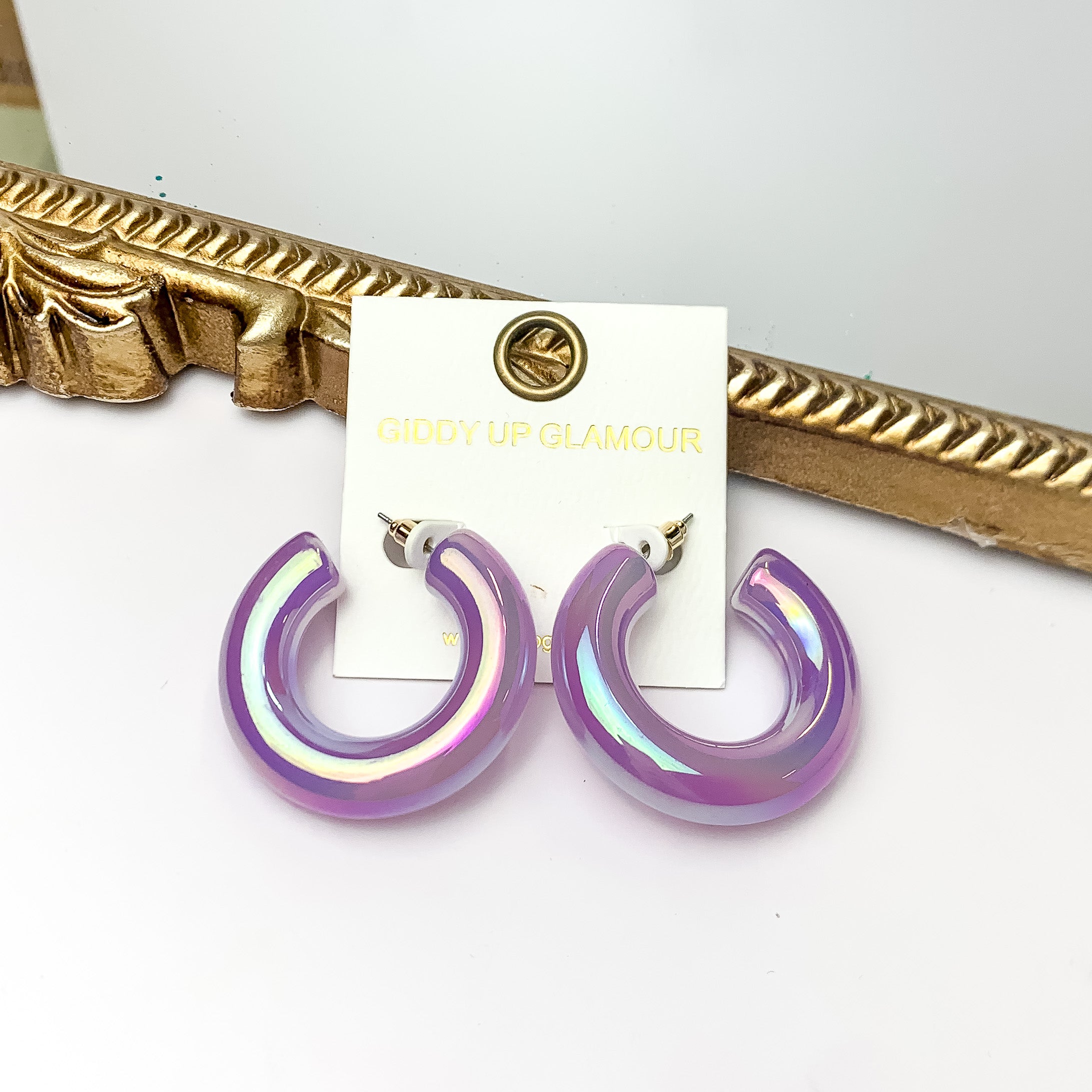 Pictured is a pair of chunky tube hoop earrings in iridescent purple. These earrings are pictured on a white background with a gold mirror behind them. 