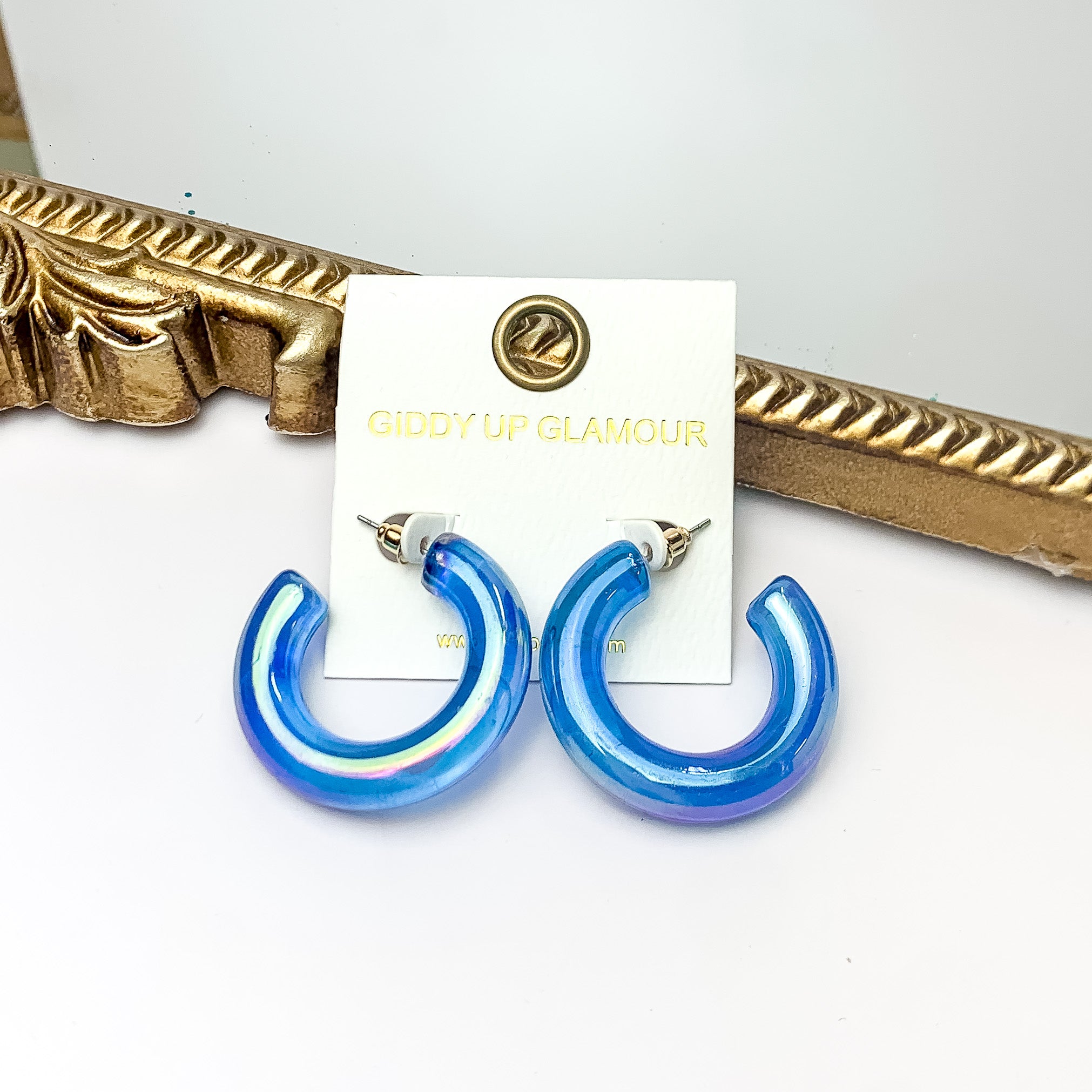 Pictured is a pair of chunky tube hoop earrings in iridescent blue. These earrings are pictured on a white background with a gold mirror behind them. 