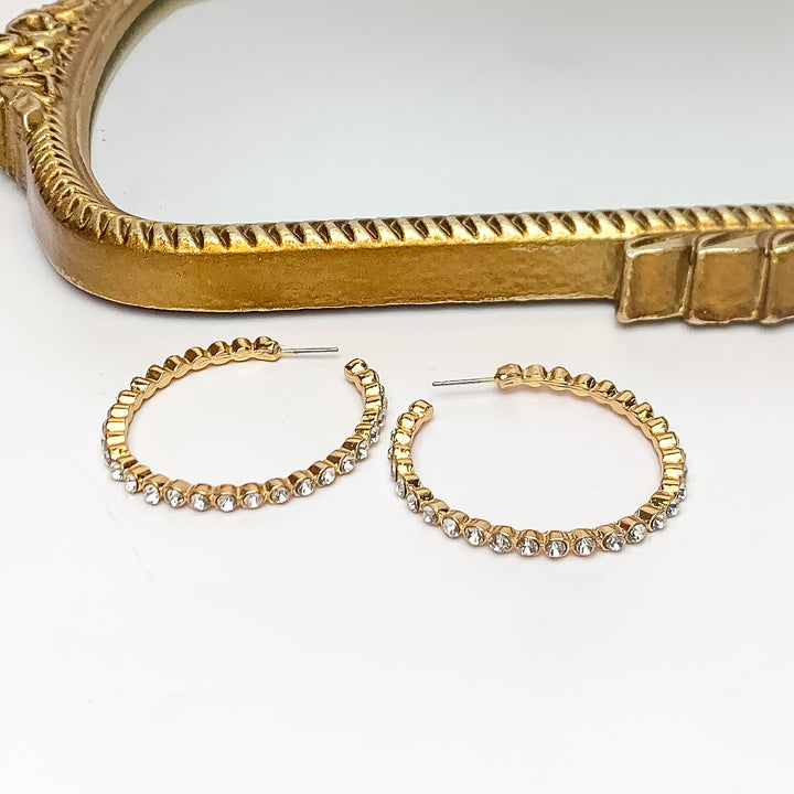 Large Gold Tone Hoop Earrings Outlined in Clear Crystals. Pictured on a white background with a gold frame above the earrings.