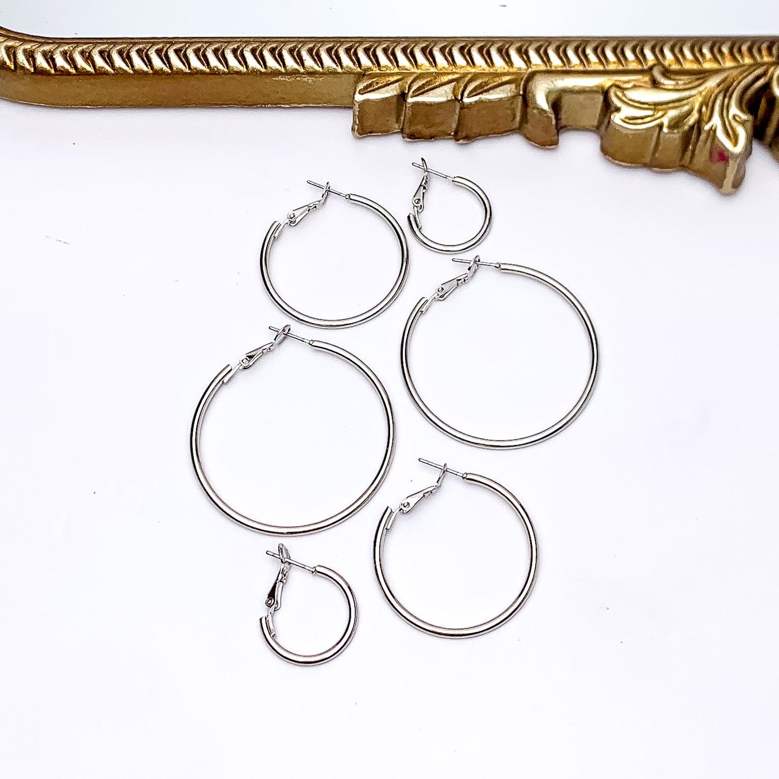 Set Of Three | Hoop Earrings in Silver Tone. Pictured on a white background with a gold frame above the earrings. There are three different sizes of earrings all in the picture.