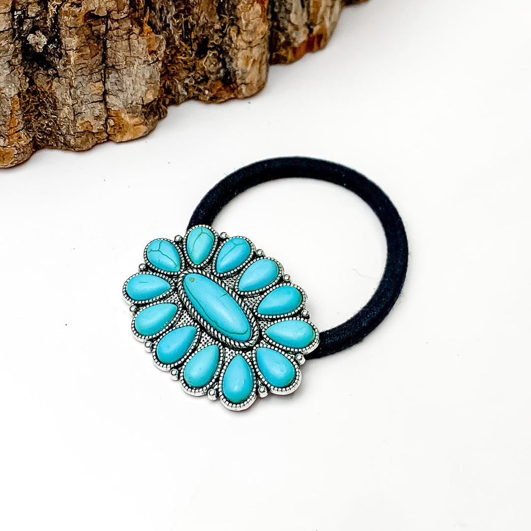Pictured is a black hair tie with a silver, oval pendant with turquoise stones. This hair tie is pictured on a white background with a piece of wood in the top left corner. 