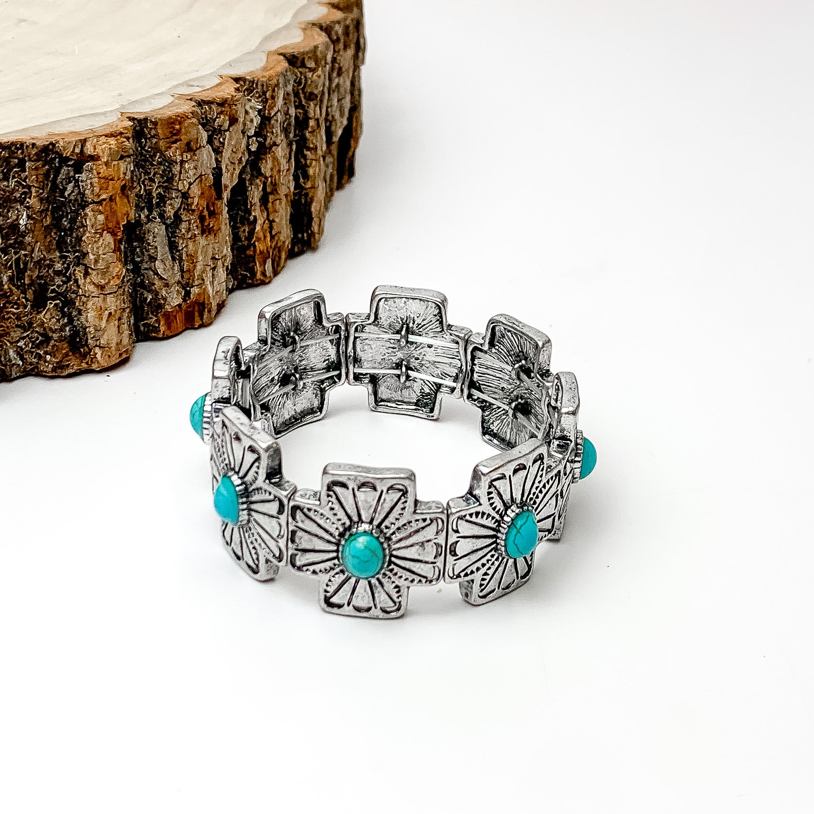 Pictured is a silver cross bracelet with engraved details and center turquoise stones. This bracelet is pictured on a white background with a piece of wood in the top left corner. 