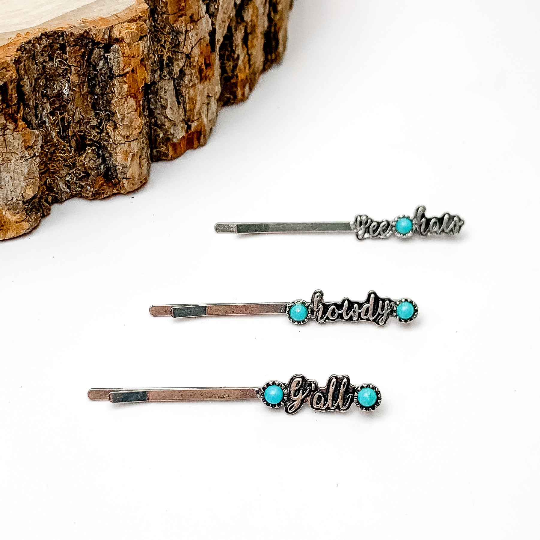 Pictured are three silver hair pins with cursive word pendants. One has the "yee-haw," one has "howdy," and one has "y'all" in cursive ad each has one or two turquoise stones. These hair pins are pictured on a white background with a piece of wood in the top left corner. 