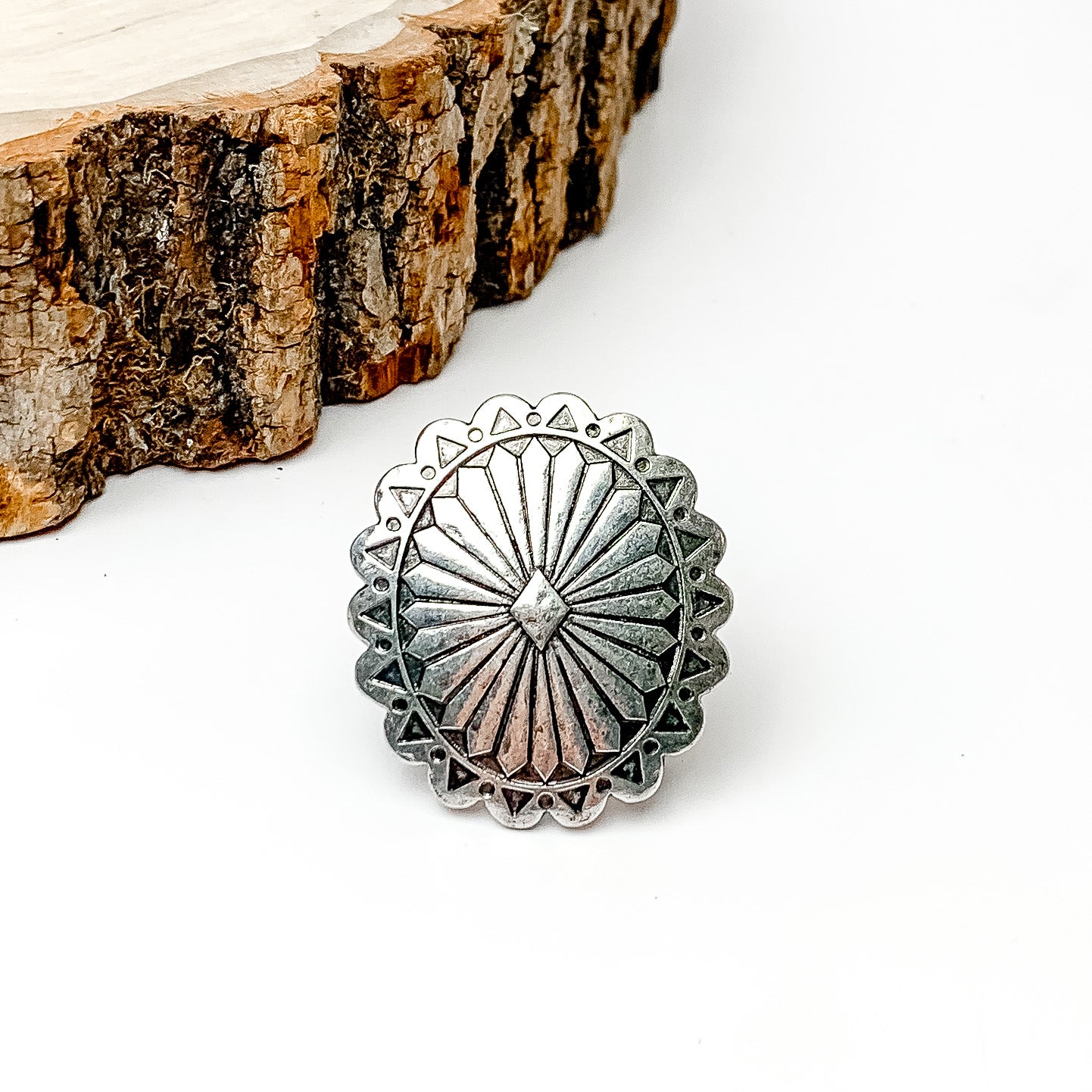 Pictured is a silver concho scarf ring. This scarf ring is pictured on a white background with a piece of wood in the top left corner. 