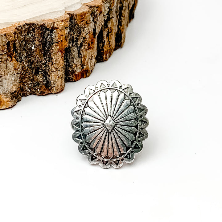 Pictured is a silver concho scarf ring. This scarf ring is pictured on a white background with a piece of wood in the top left corner. 