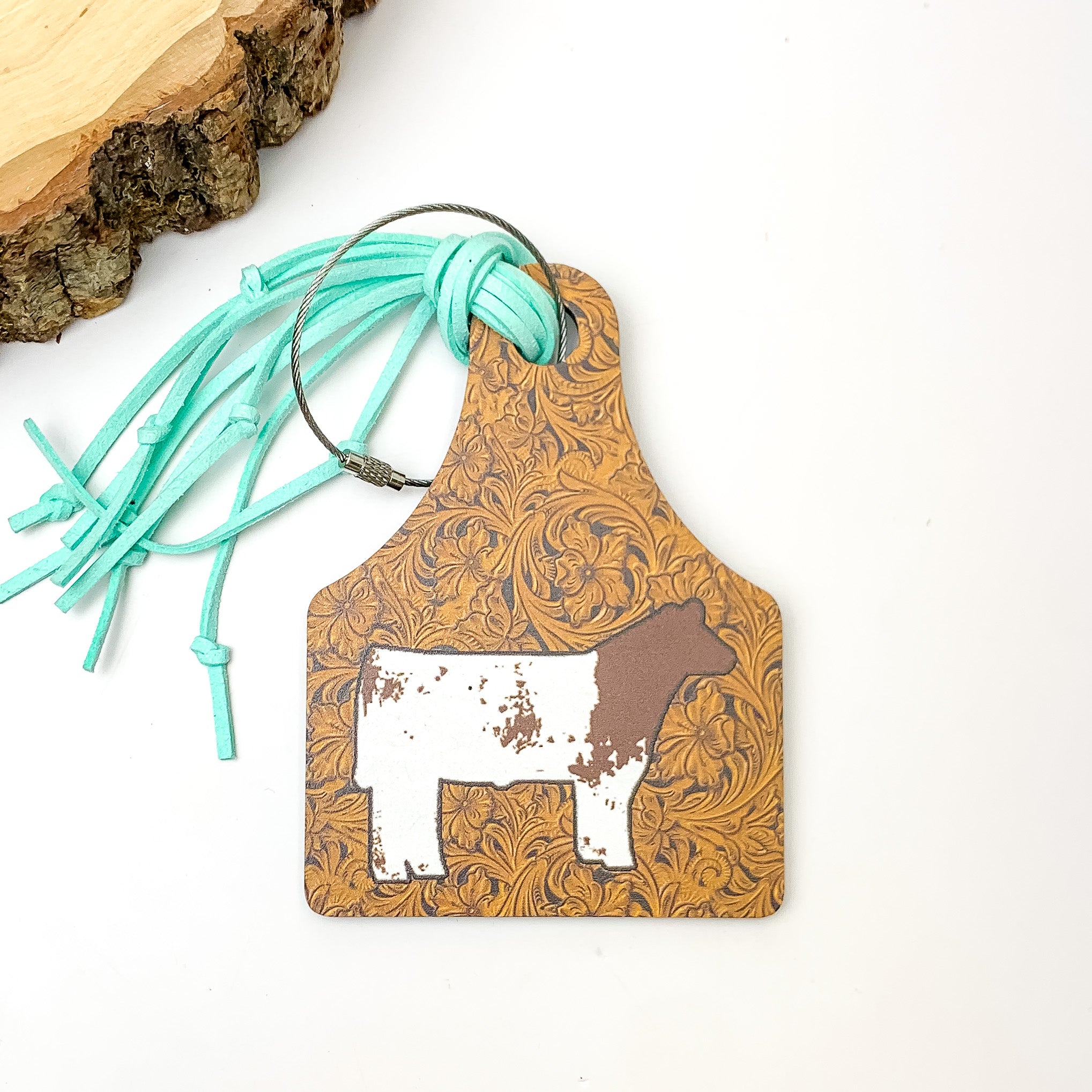 Brown and Turquoise Luggage Tag With Cow. Pictured on a white background with a wood piece in the top left corner.