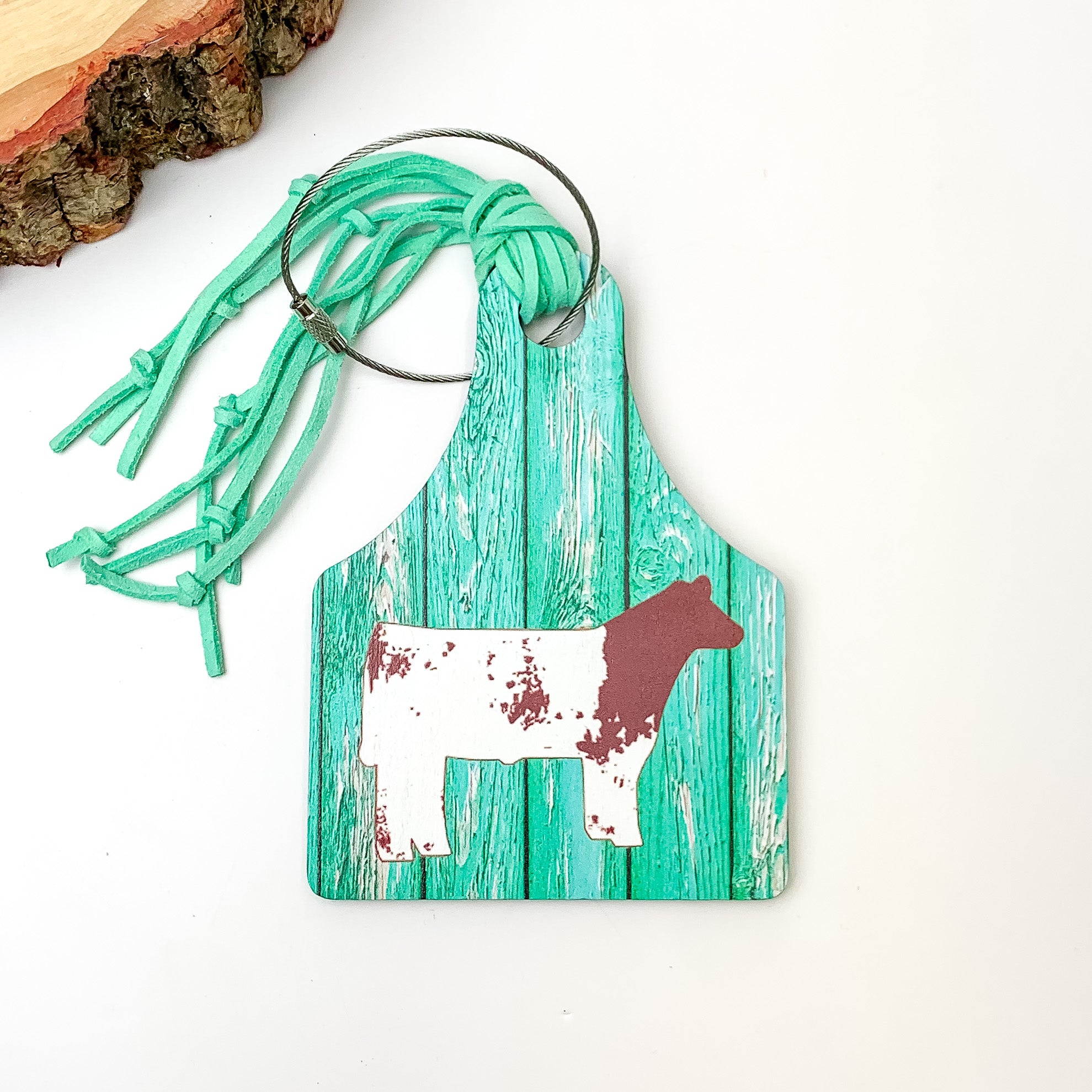 Turquoise Luggage Tag With Cow. Pictured on a white background with a wood piece in the top left corner.