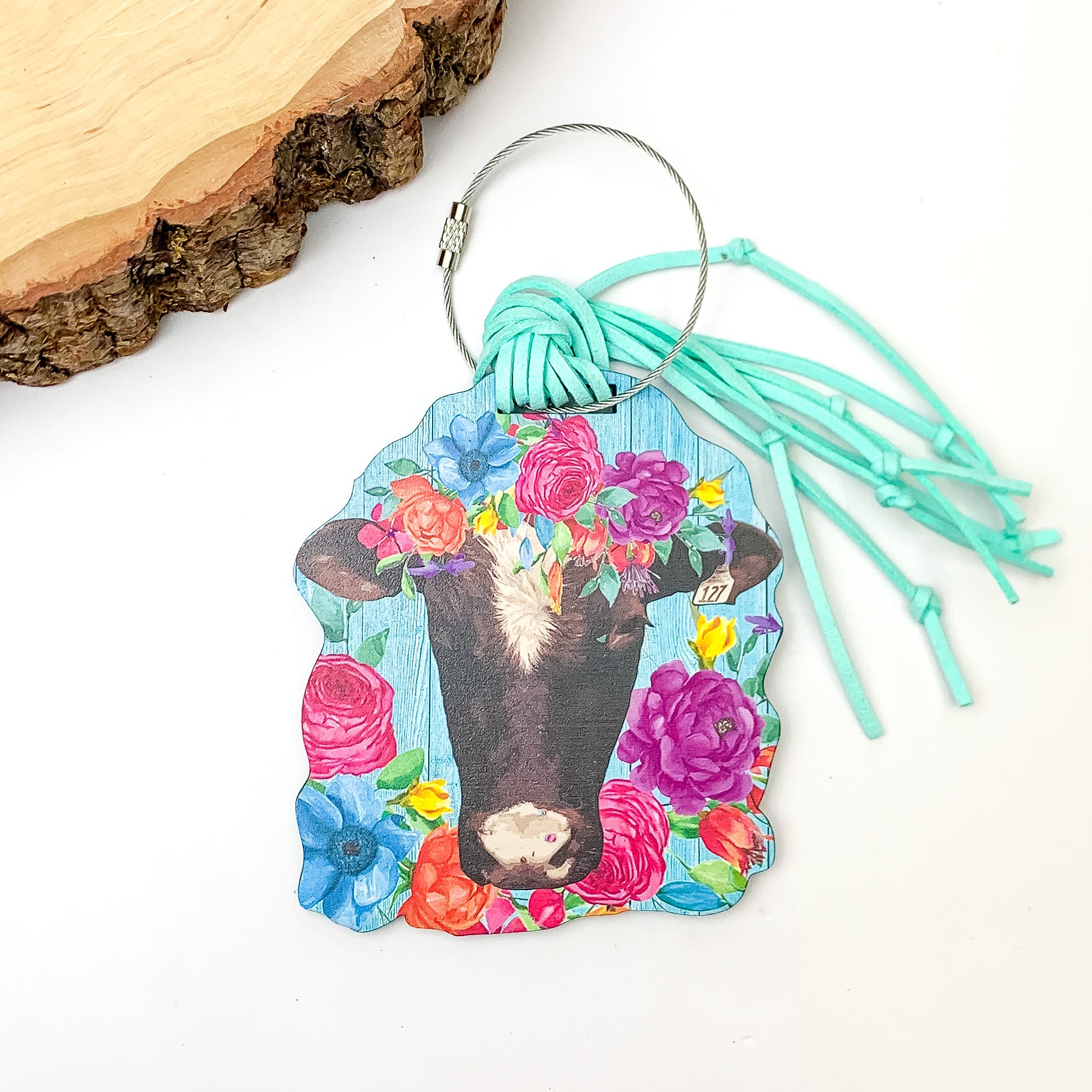 Cow Head Luggage Tag With Flower Crown in Multicolor. Pictured on a white background with a wood piece in the top left corner.
