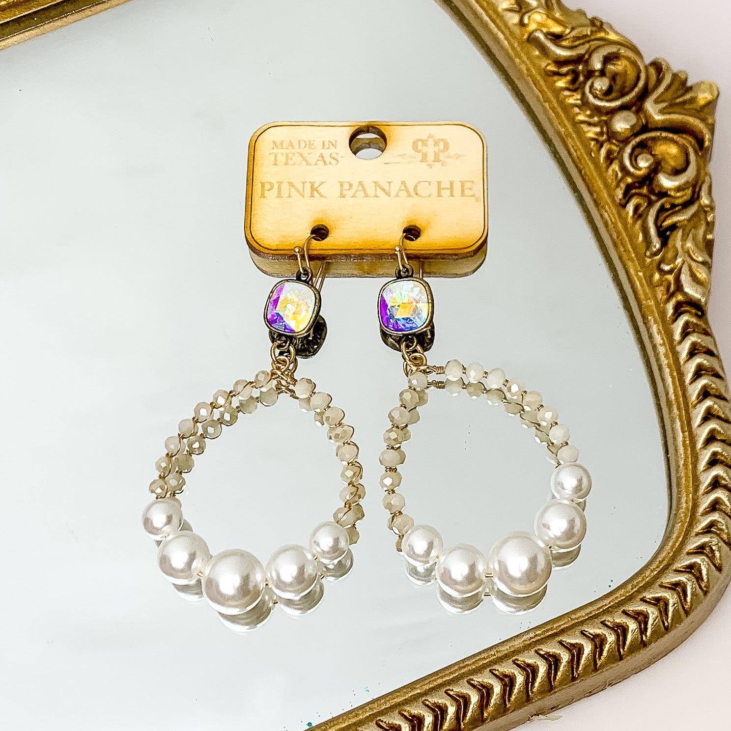 AB cushion cut on gold fish hook earrings with a white crystal and white pearl beaded open circle. These earrings are pictured on a wooden earrings holder on a gold mirror. 