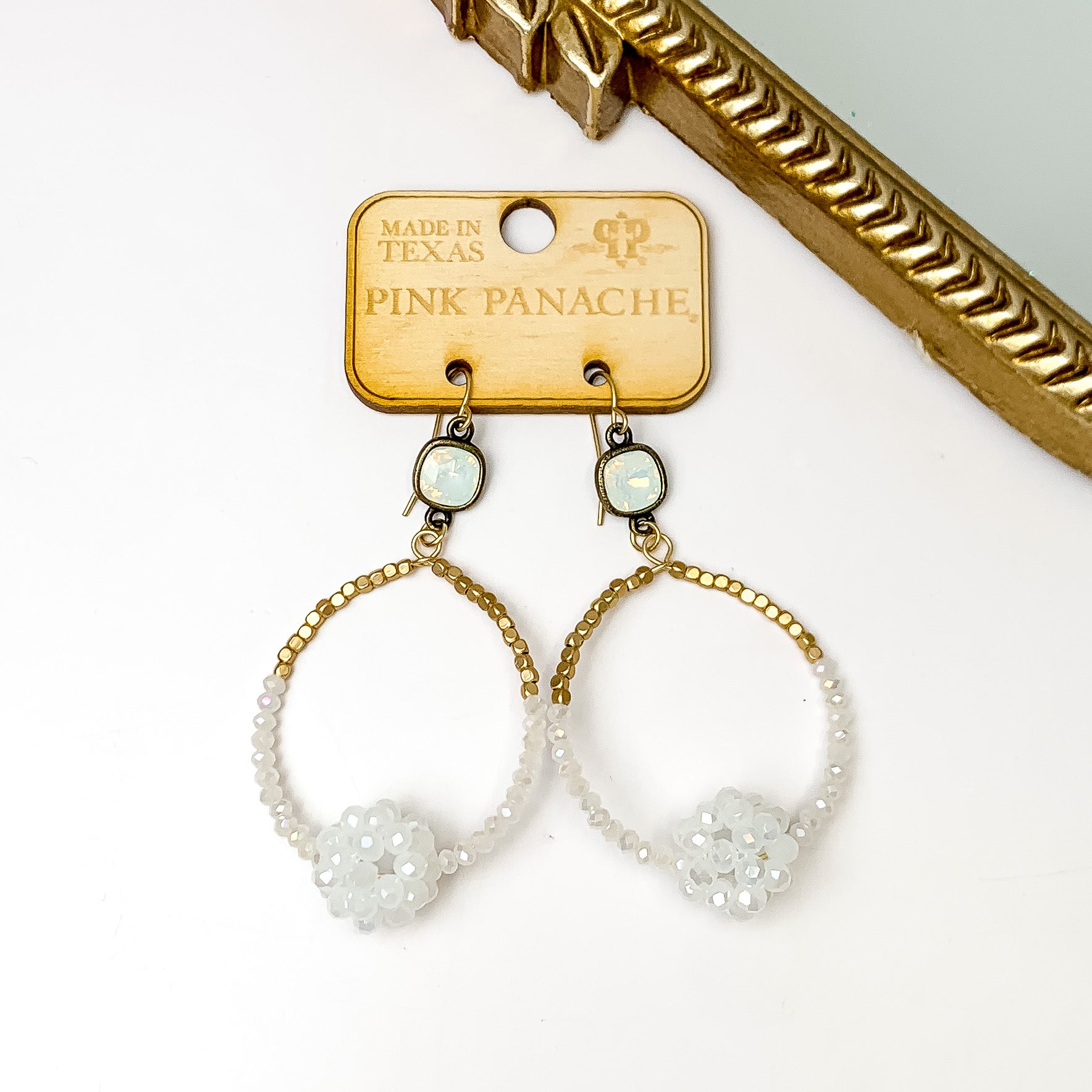 White opal cushion cut on gold fish hook earrings with a gold and white crystal beaded open circle. These earrings are pictured on a wooden earrings holder on a white background with a gold mirror in the top right corner. 