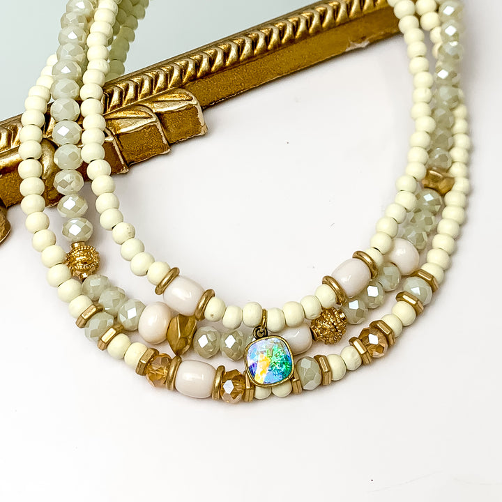 Pink Panache Triple Strand Ivory Crystal and Wood Beaded Necklace With Bronze/ AB Cushion Cut Drop. Pictured on a white background with the top of the necklace on a mirror with a gold trim.