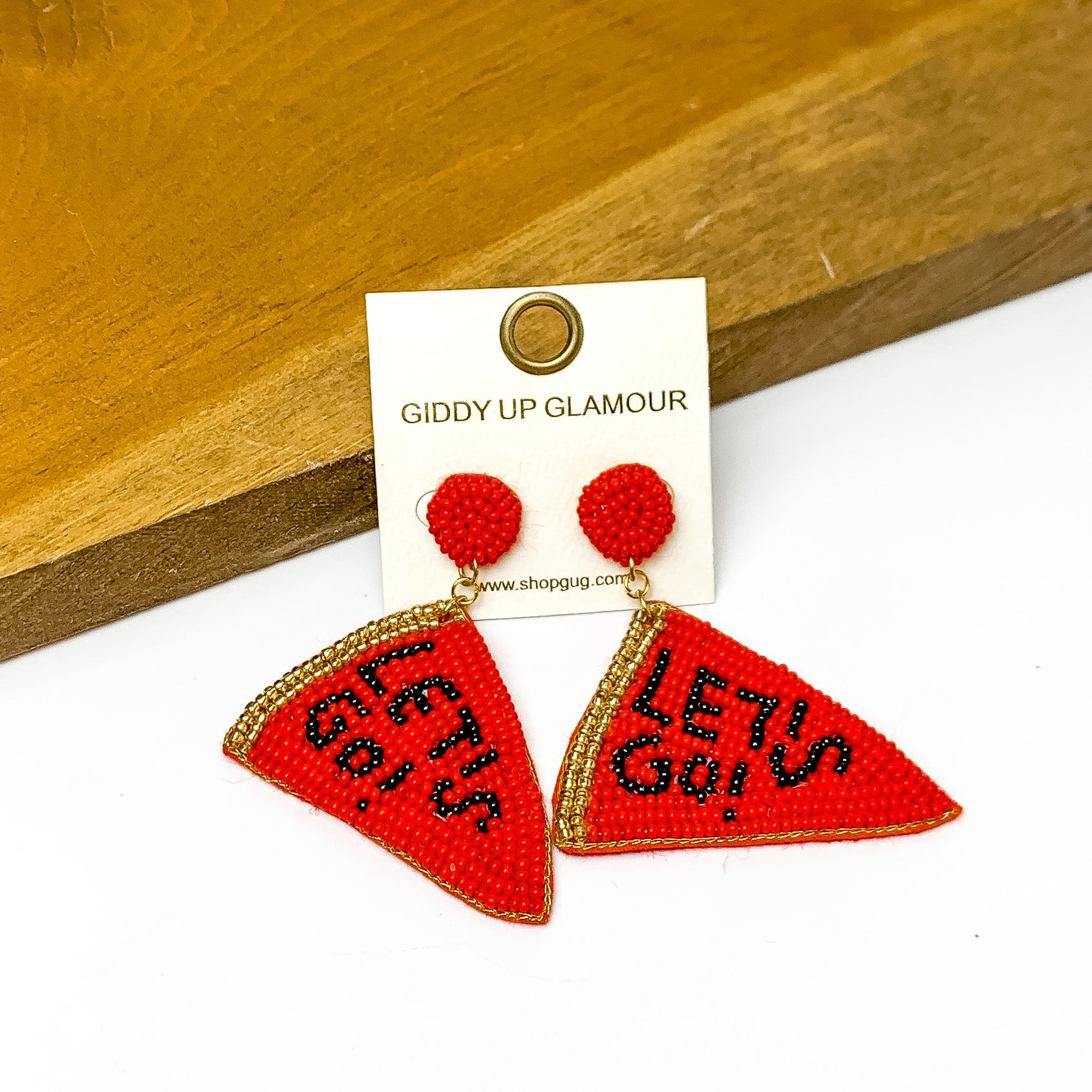 Lets Go Beaded Flag Earrings in Red. Pictured on a white background with the earrings laying against a wood piece. 