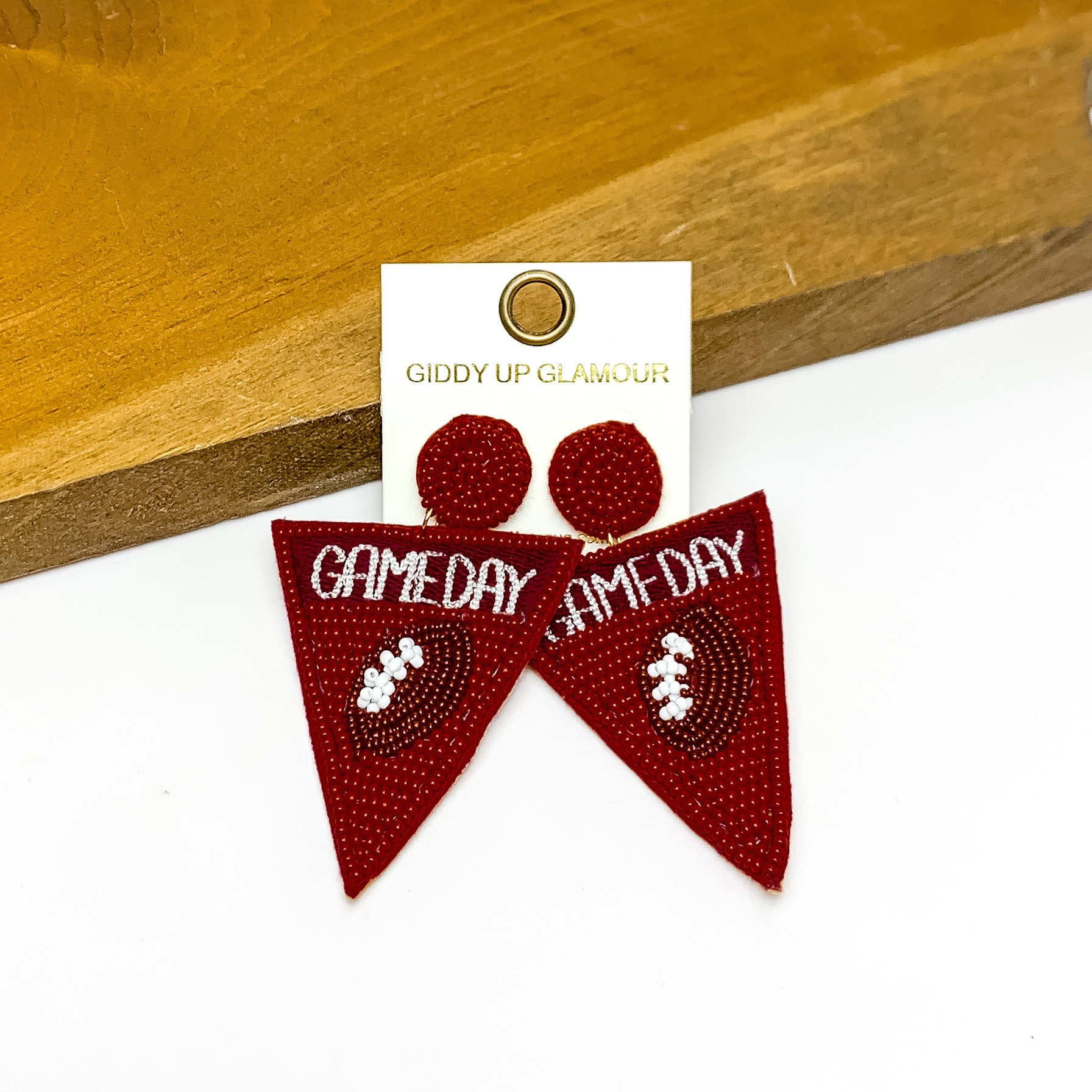 Gameday Beaded Flag Earrings in Maroon. Pictured on a white background with the earrings laying against a piece of wood.. 