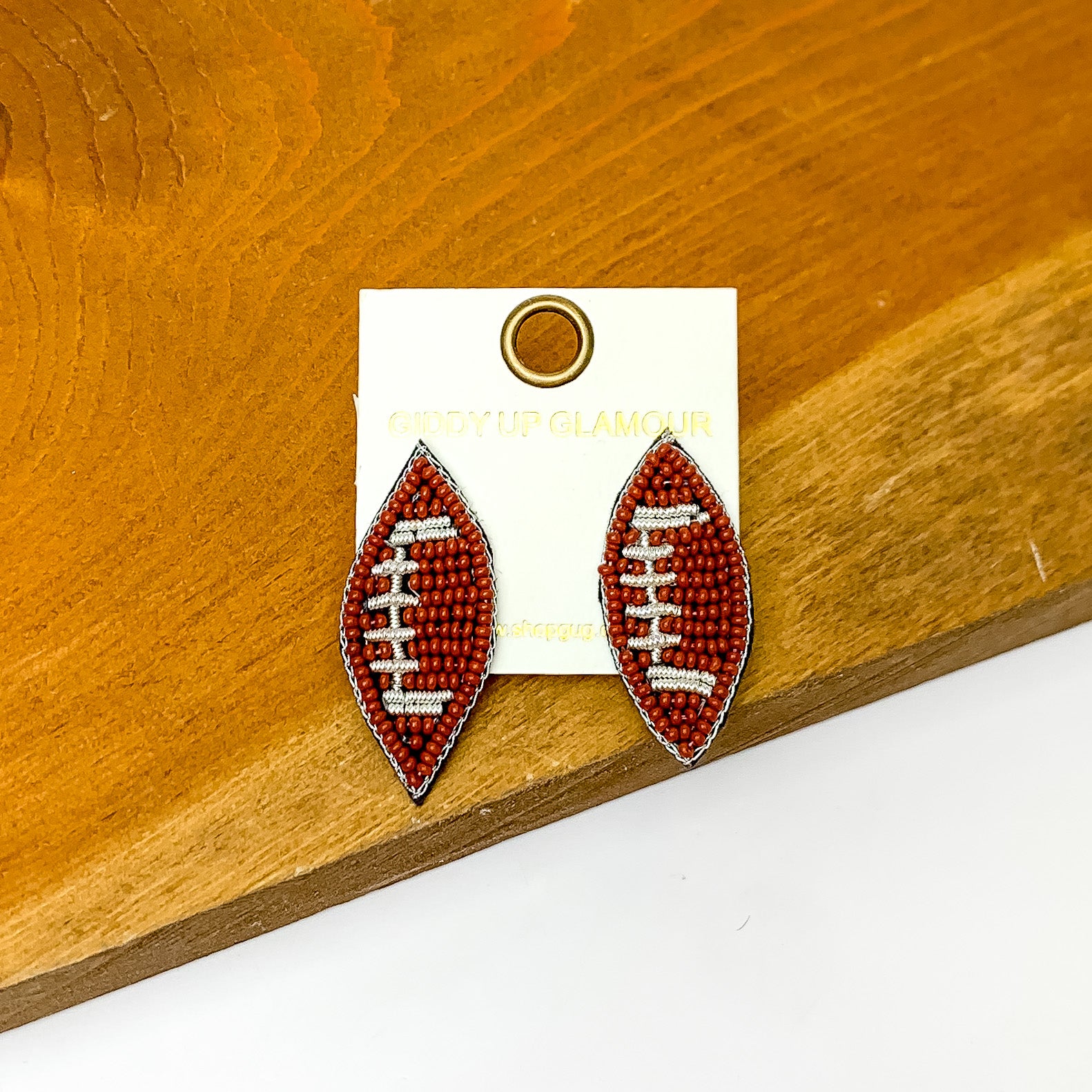 Let's Get Sporty Seed Bead Football Stud Earrings. Pictured on a white background with the earrings laying on a wood piece.