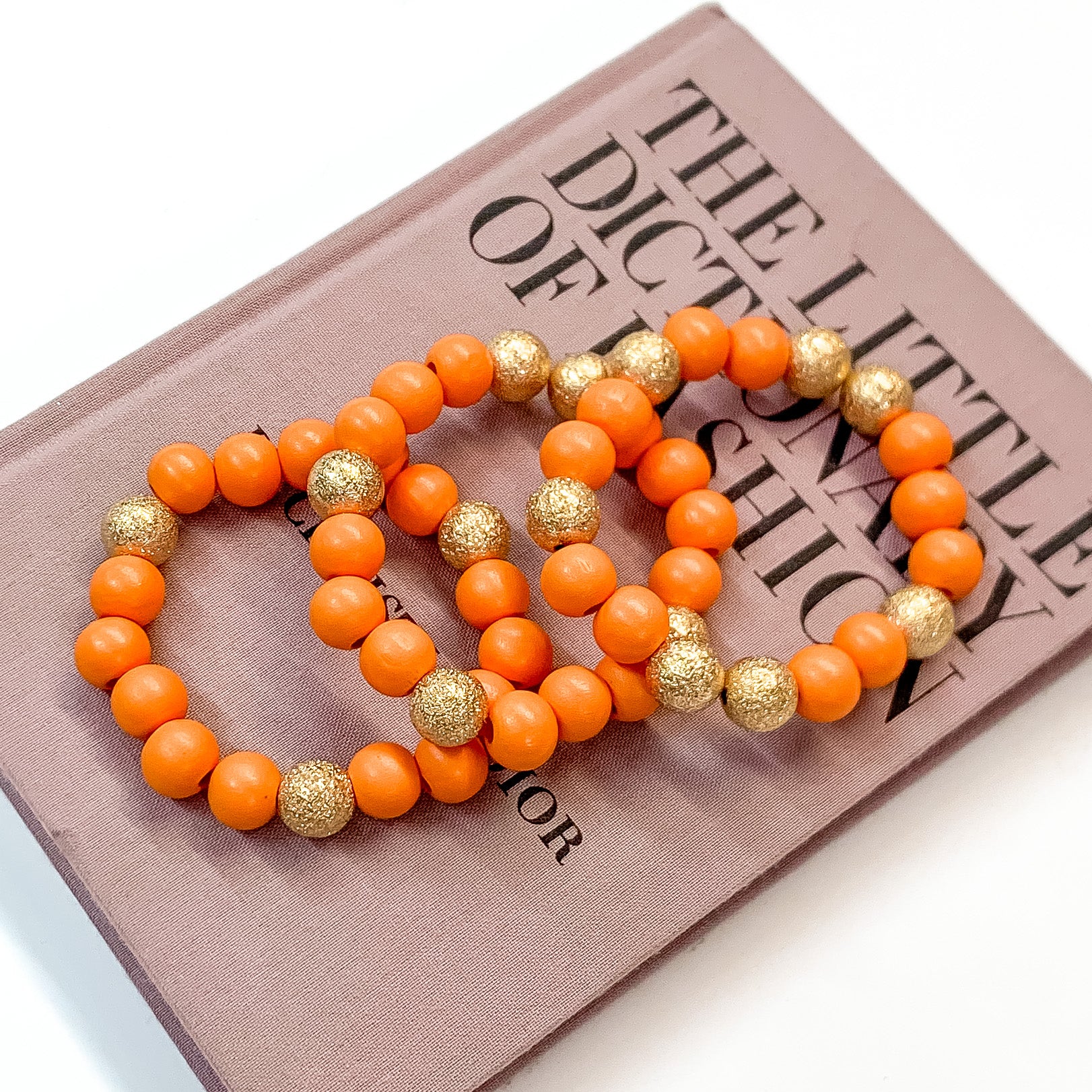 Pictured on a mauve colored book is a set of orange beaded bracelets with gold beaded spacers. 