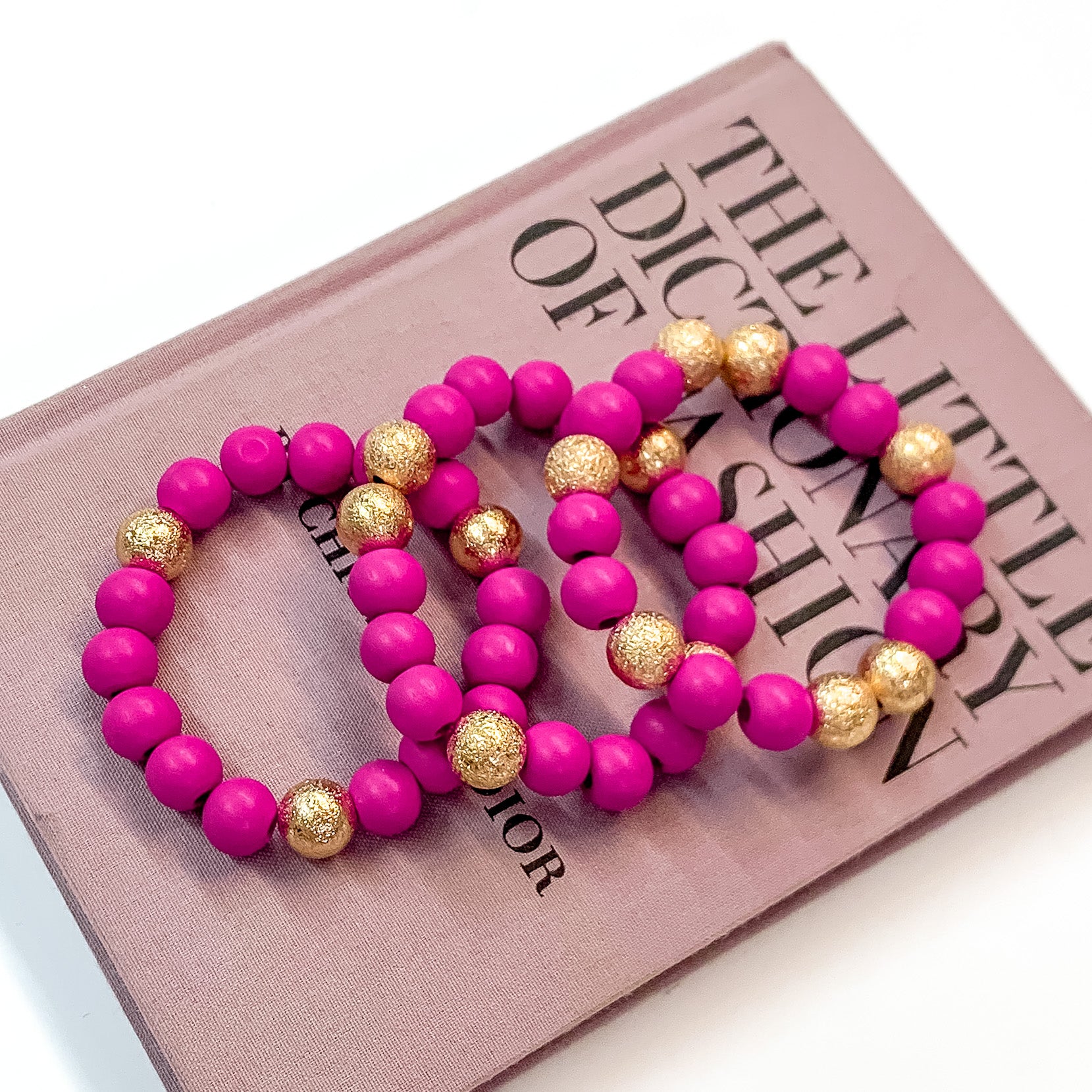 Pictured on a mauve colored book is a set of fuchsia purple beaded bracelets with gold beaded spacers.