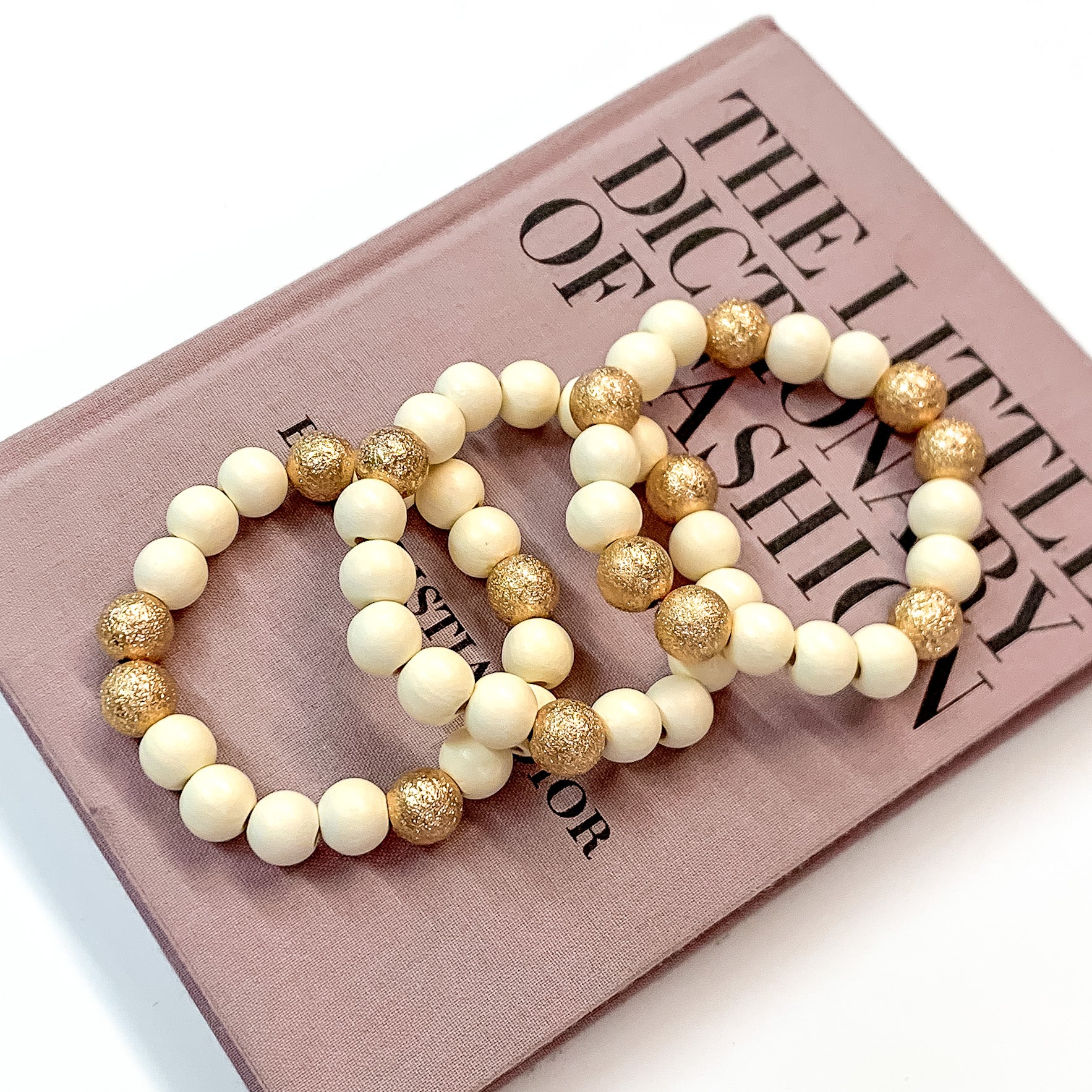 Pictured on a mauve colored book is a set of ivory beaded bracelets with gold beaded spacers. 