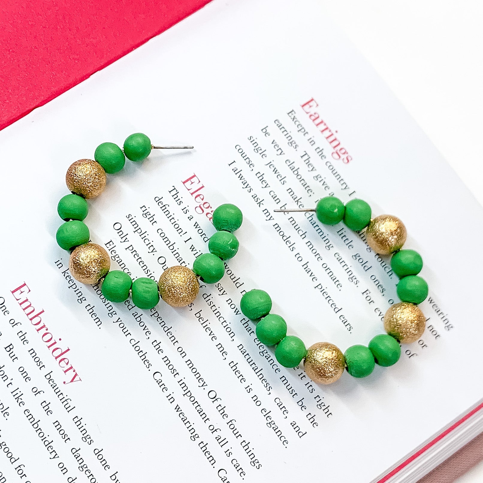 Pictured on an open book is a pair of green beaded hoop earrings with gold beaded spacers. 