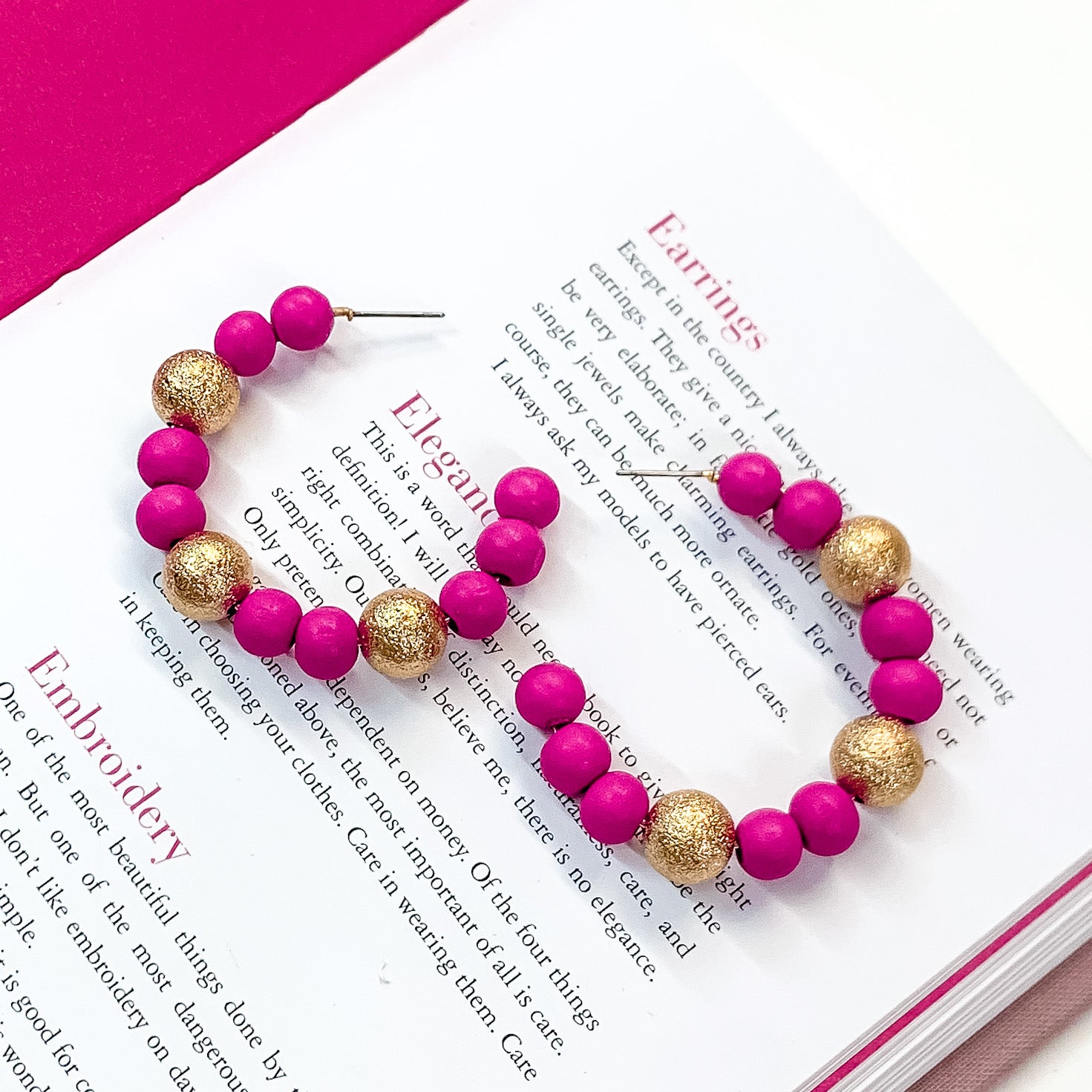 Pictured on an open book is a pair of fuchsia beaded hoop earrings with gold beaded spacers.