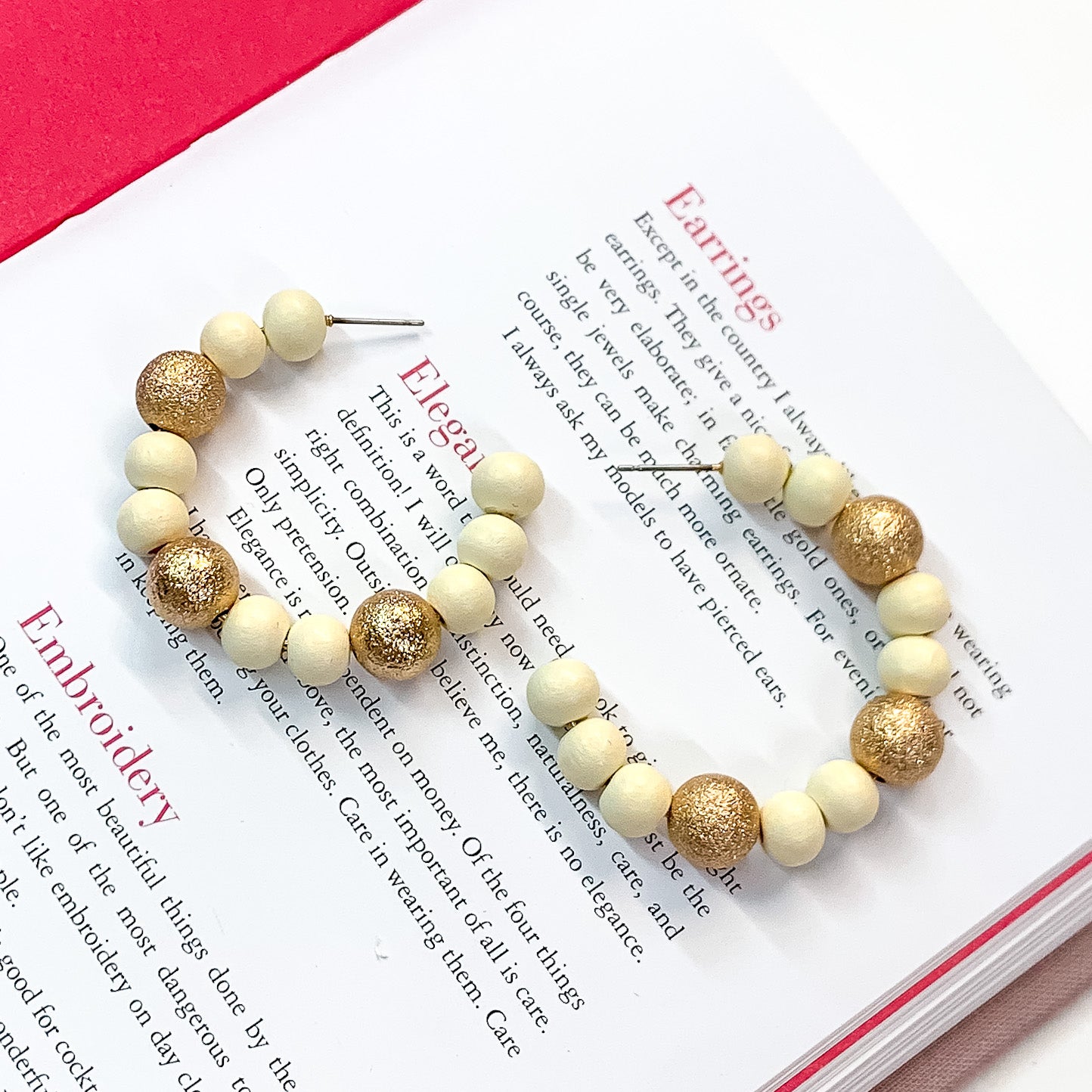 Pictured on an open book is a pair of ivory beaded hoop earrings with gold beaded spacers. 