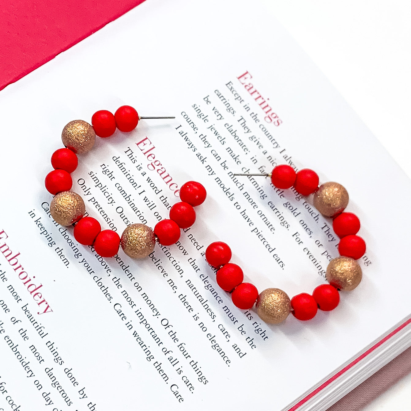 Pictured on an open book is a pair of red beaded hoop earrings with gold beaded spacers. 