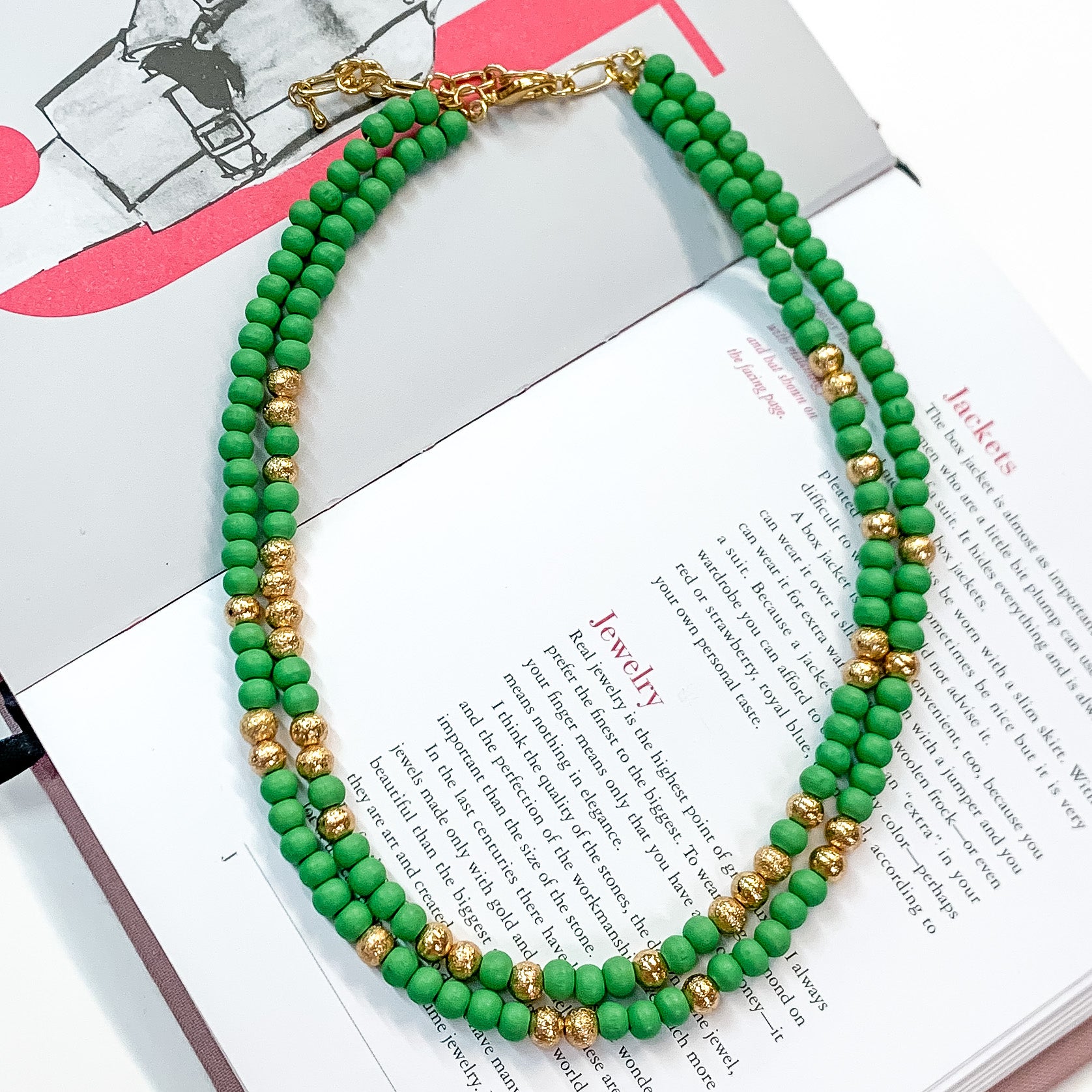 Pictured on an open book is a two strand green beaded necklace with gold beaded spacers and gold hardware. 