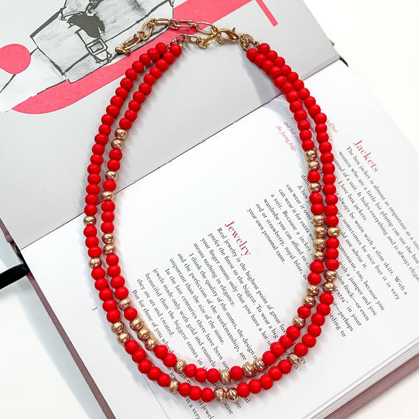 Pictured on an open book is a two strand red beaded necklace with gold beaded spacers and gold hardware. 