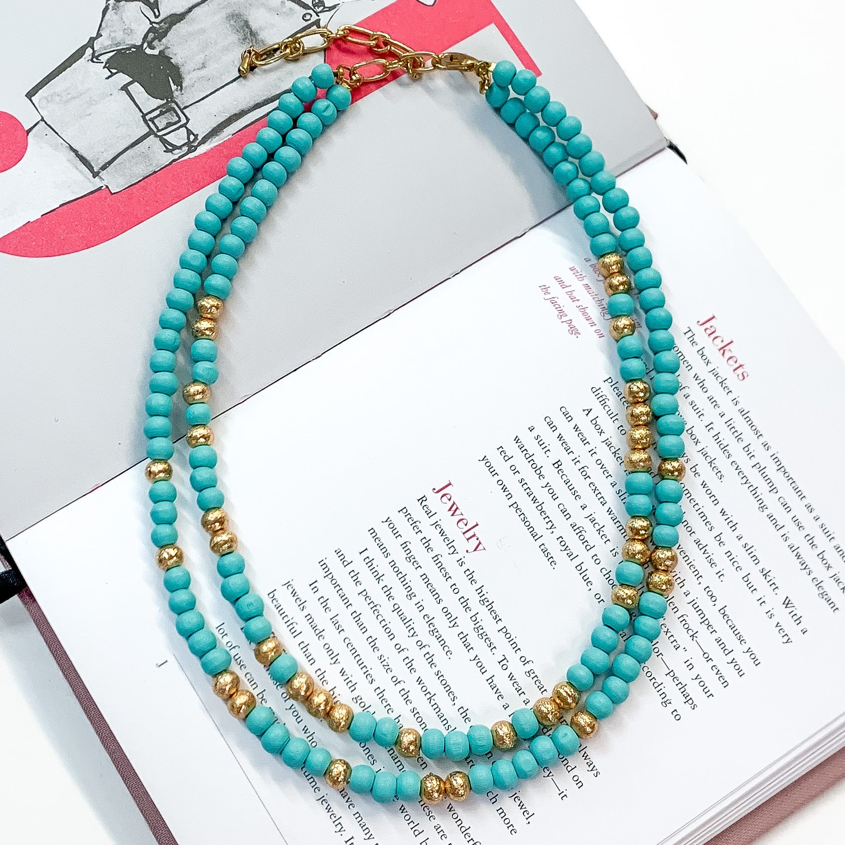 Pictured on an open book is a two strand sky blue beaded necklace with gold beaded spacers and gold hardware. 