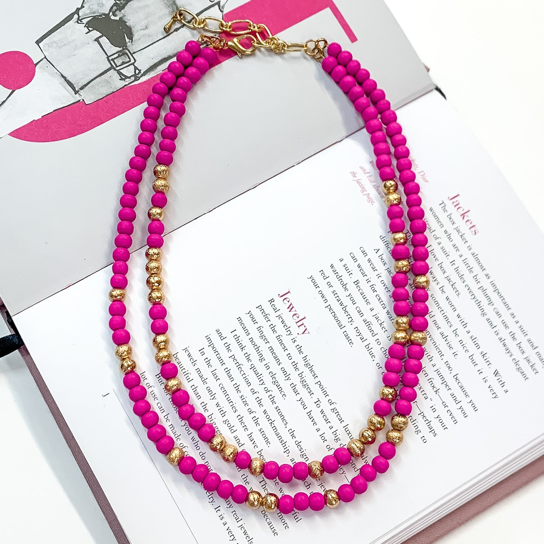 Pictured on an open book is a two strand fuchsia beaded necklace with gold beaded spacers and gold hardware.