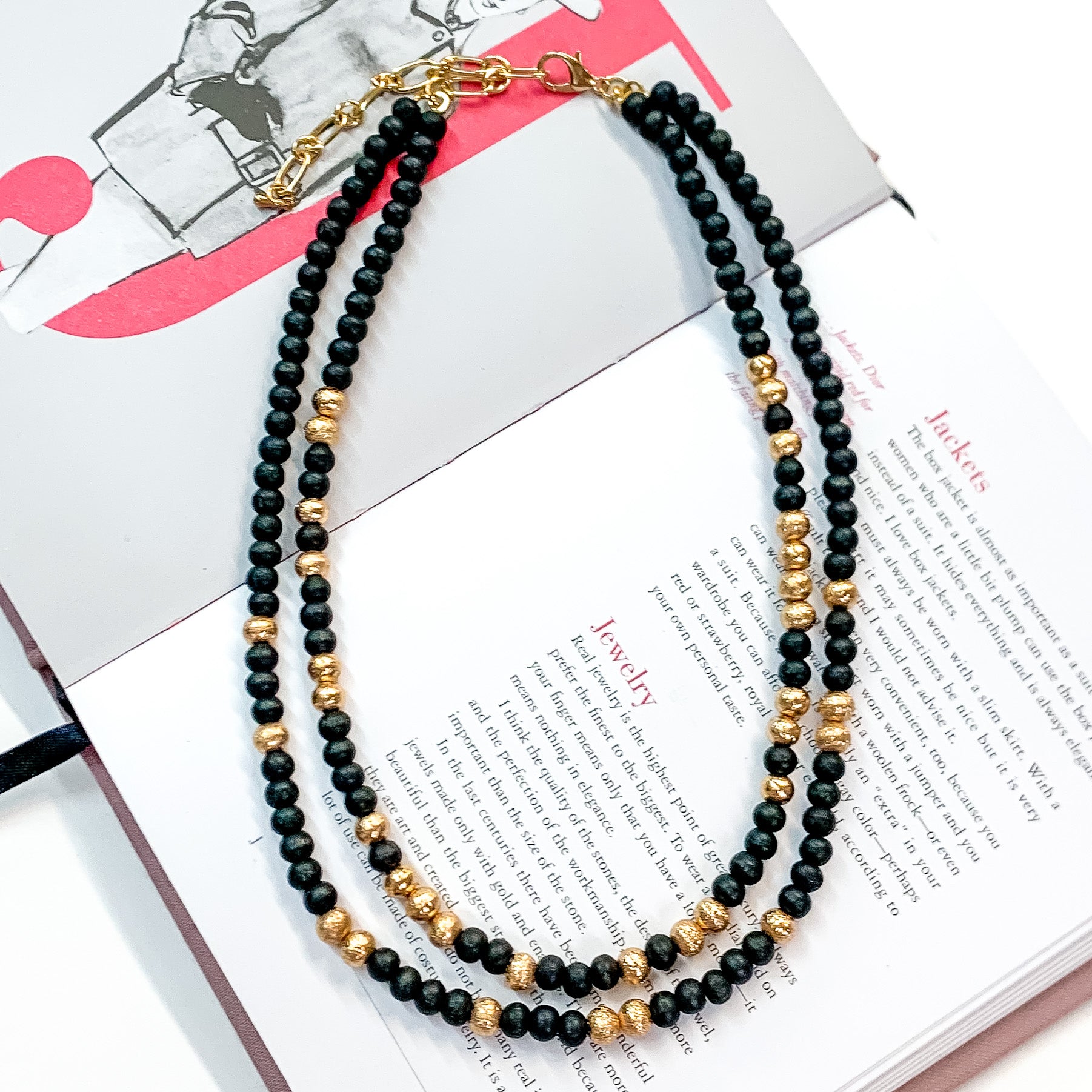 Pictured on an open book is a two strand black beaded necklace with gold beaded spacers and gold hardware. 