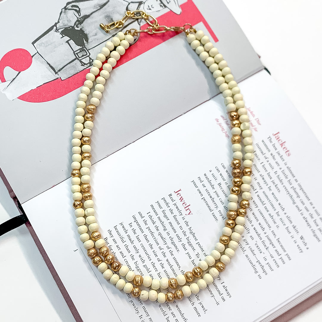 Pictured on an open book is a two strand ivory beaded necklace with gold beaded spacers and gold hardware. 
