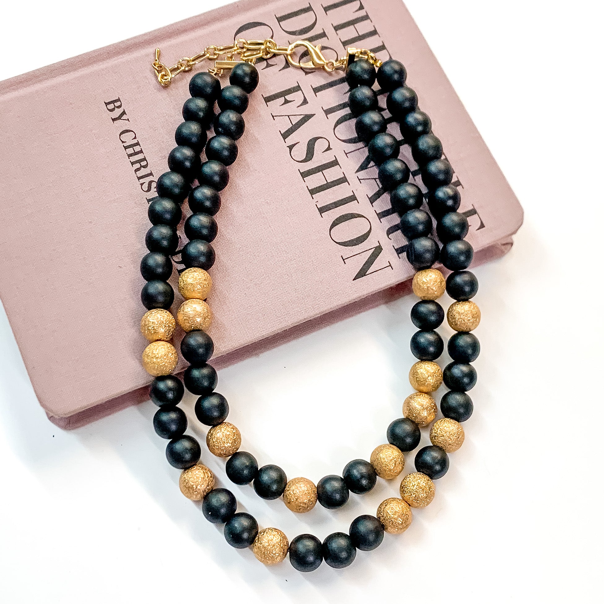 Pictured partially on a mauve colored book on a white background is a two strand black beaded necklace with gold beaded spacers and gold hardware. 