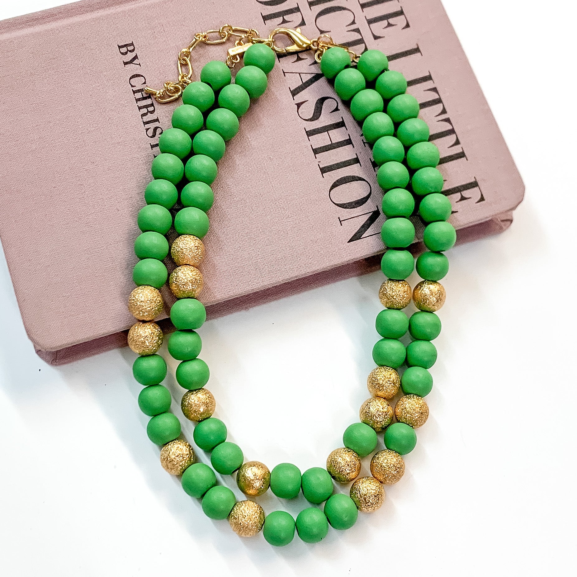 Pictured partially on a mauve colored book on a white background is a two strand green beaded necklace with gold beaded spacers and gold hardware. 