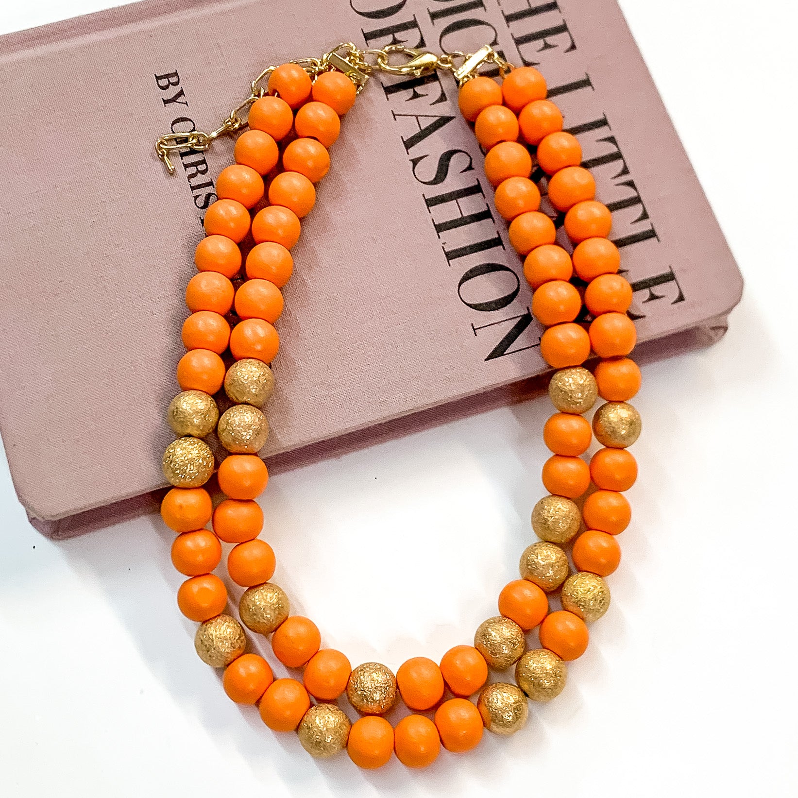 Pictured partially on a mauve colored book on a white background is a two strand orange beaded necklace with gold beaded spacers and gold hardware. 
