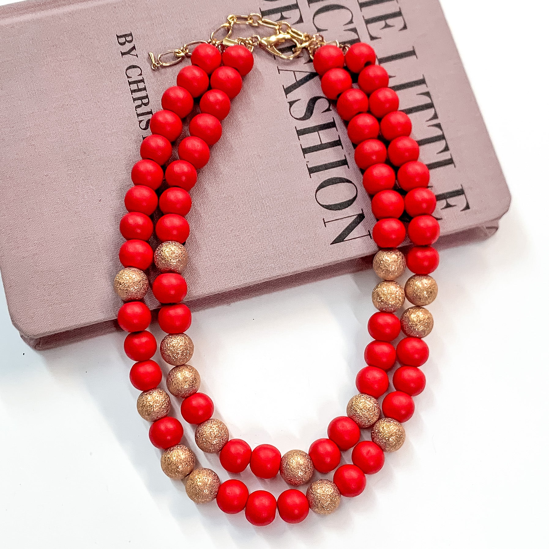 Pictured partially on a mauve colored book on a white background is a two strand red beaded necklace with gold beaded spacers and gold hardware. 