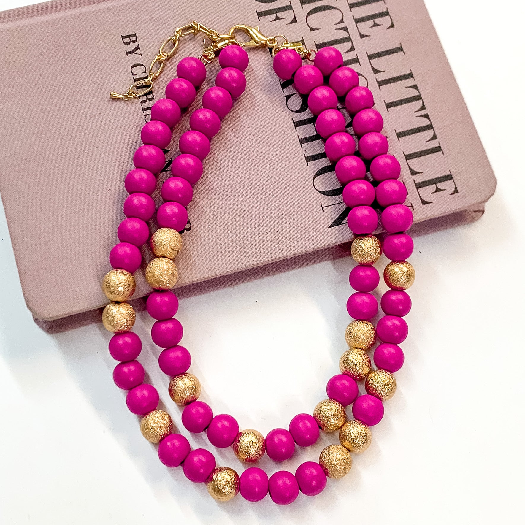 Pictured partially on a mauve colored book on a white background is a two strand fuchsia beaded necklace with gold beaded spacers and gold hardware. 