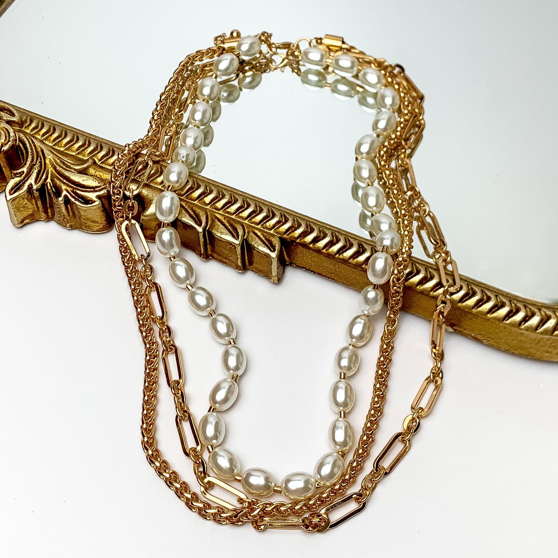Princess Pearl Three Strand Necklace in Gold Tone. Pictured on a white background with part of the necklace laying on a mirror with a gold trim.