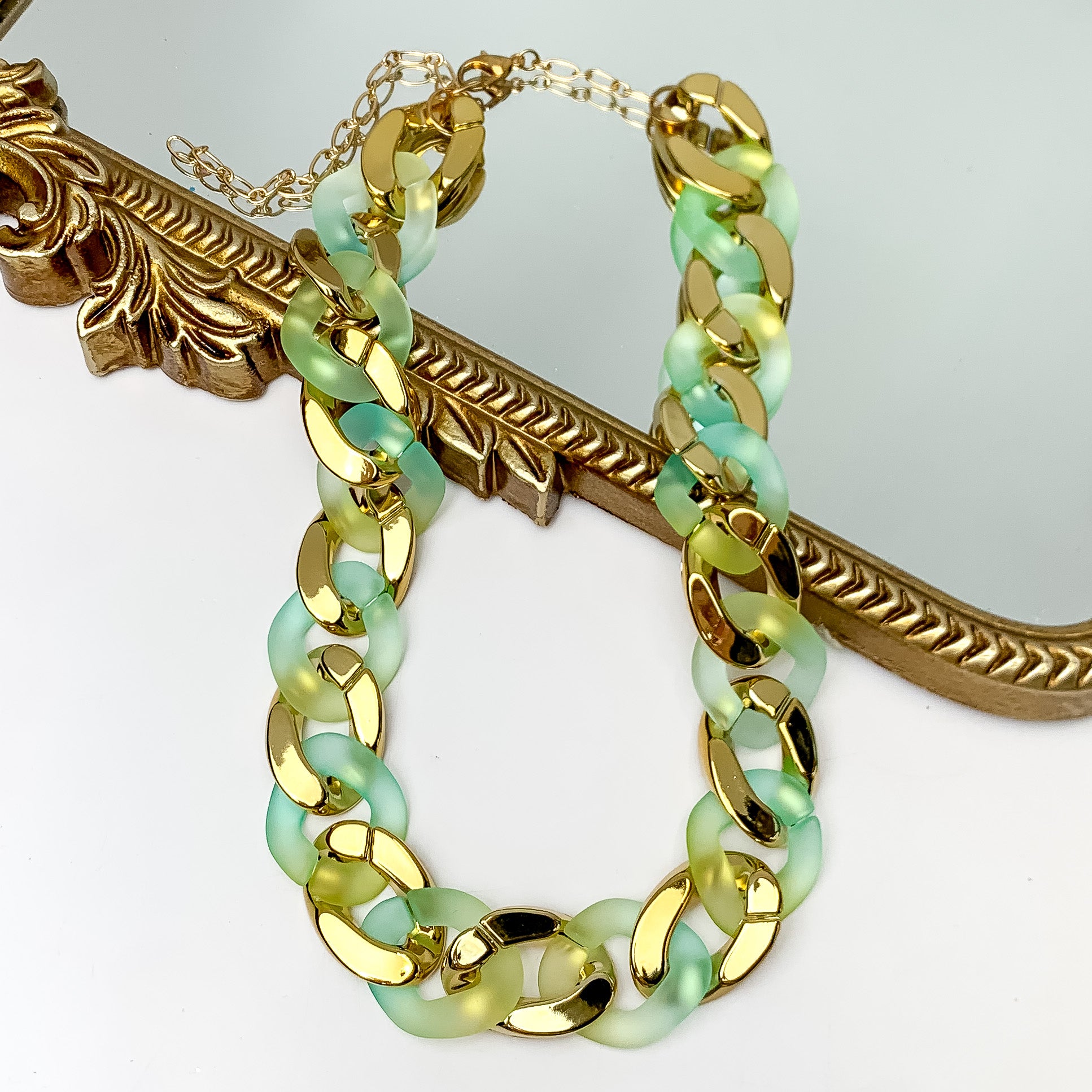 Pictured partially on a gold mirror on a white background is a chain link necklace in gold and green. 